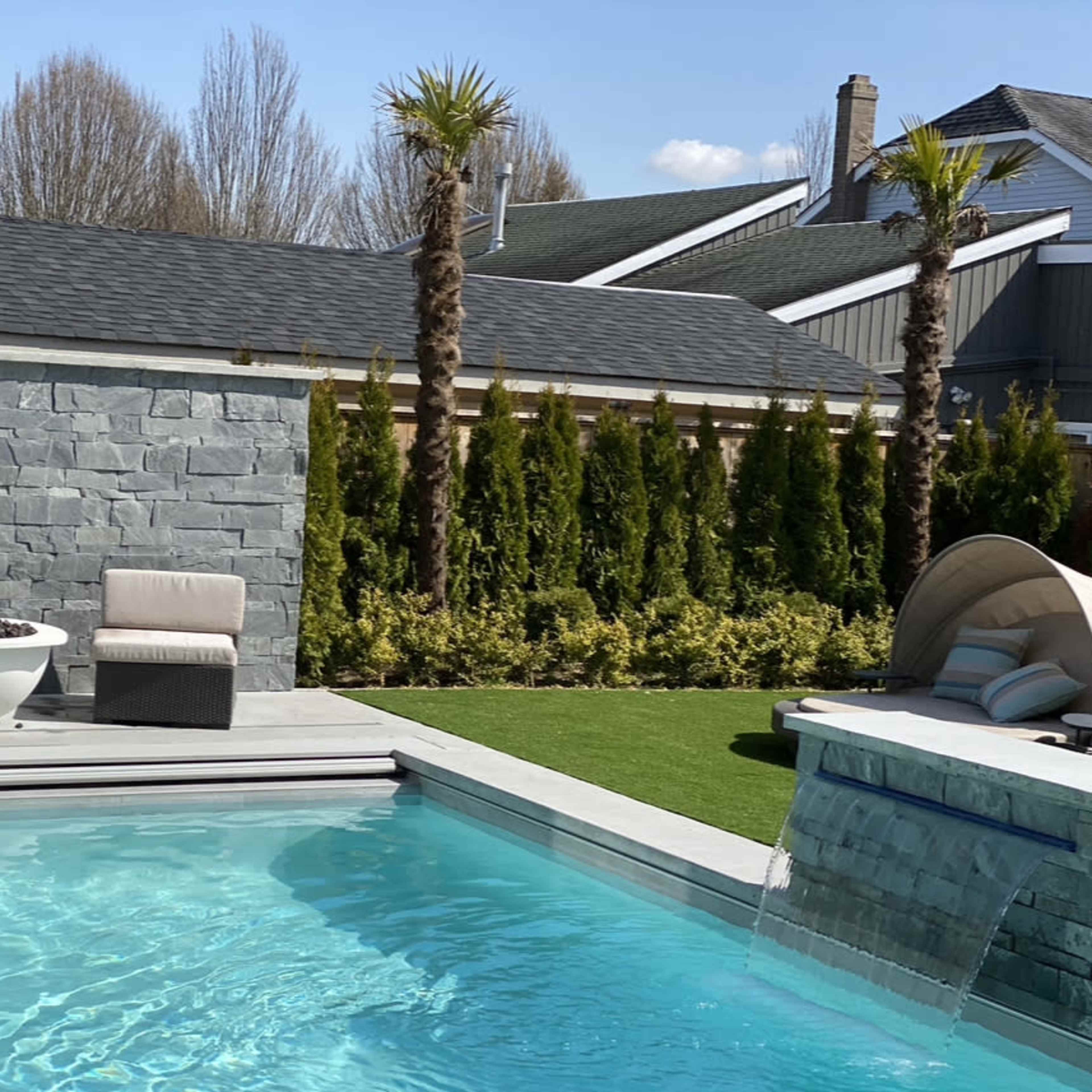 A modern swimming pool with a waterfall feature, surrounded by lounge chairs and palm trees, set against a backdrop of landscaped hedges and buildings.