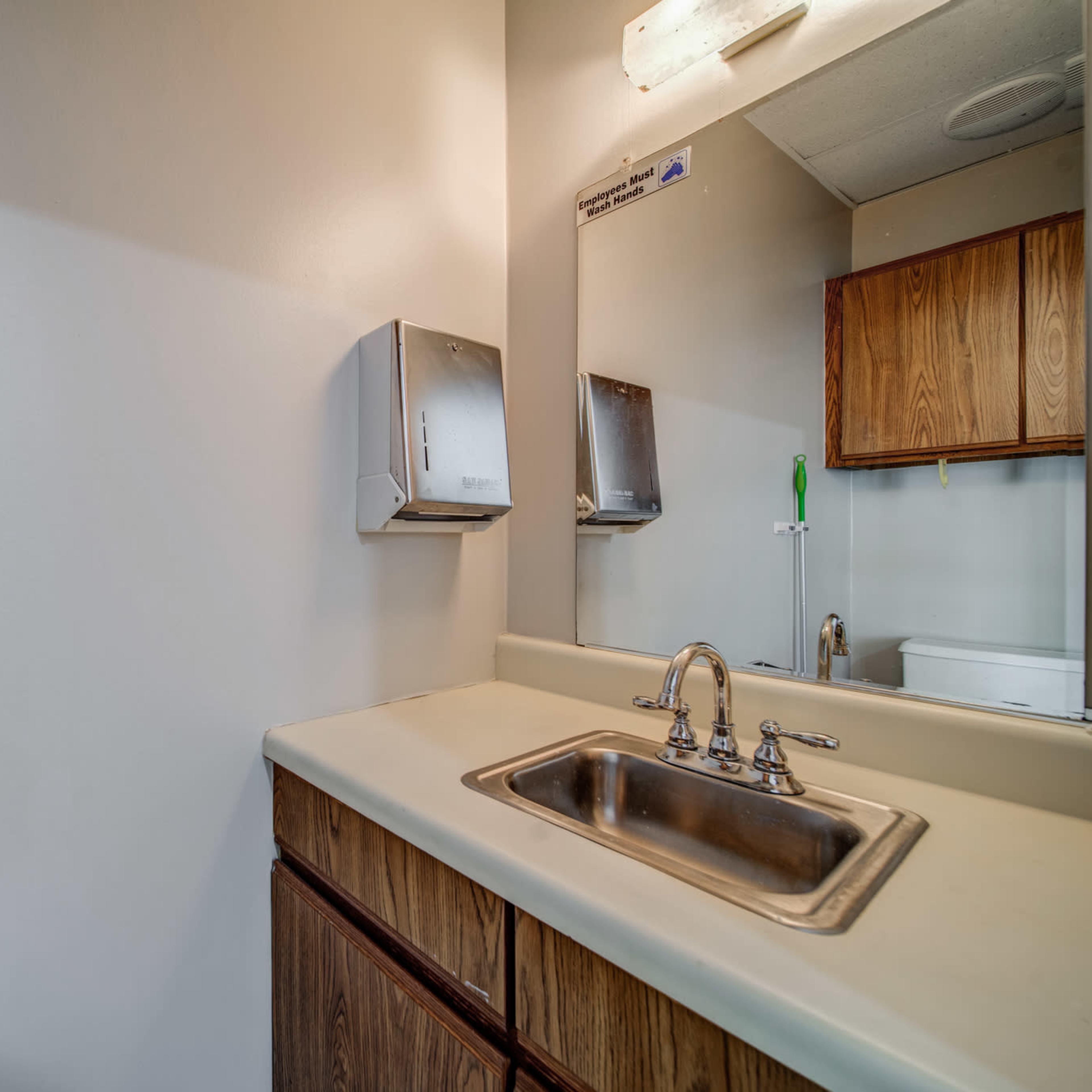 A small restroom with a sink, mirror, and wooden cabinets, alongside a toilet visible in the background.