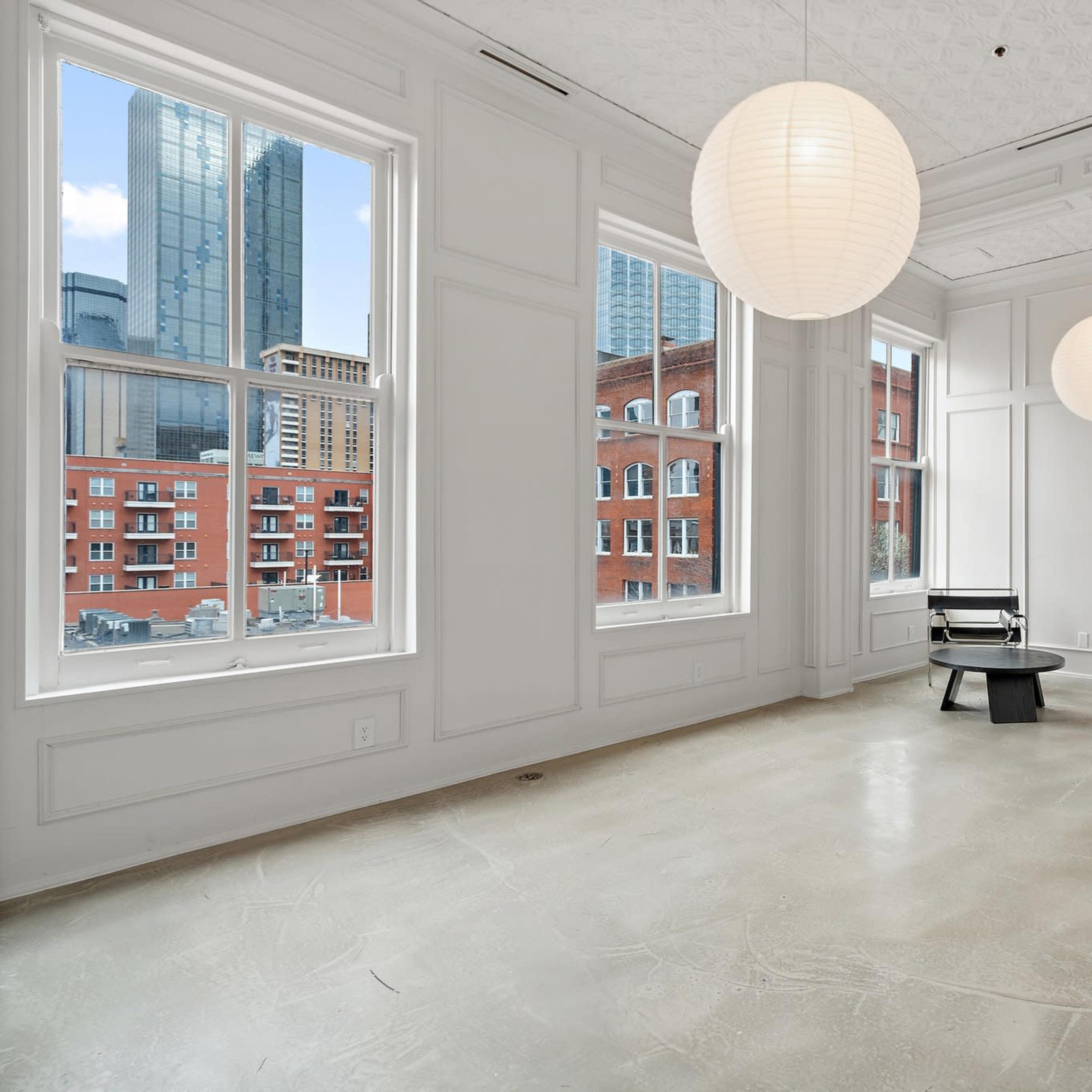 A spacious, minimalist room with large windows, two round hanging lamps, and a single chair on a concrete floor.