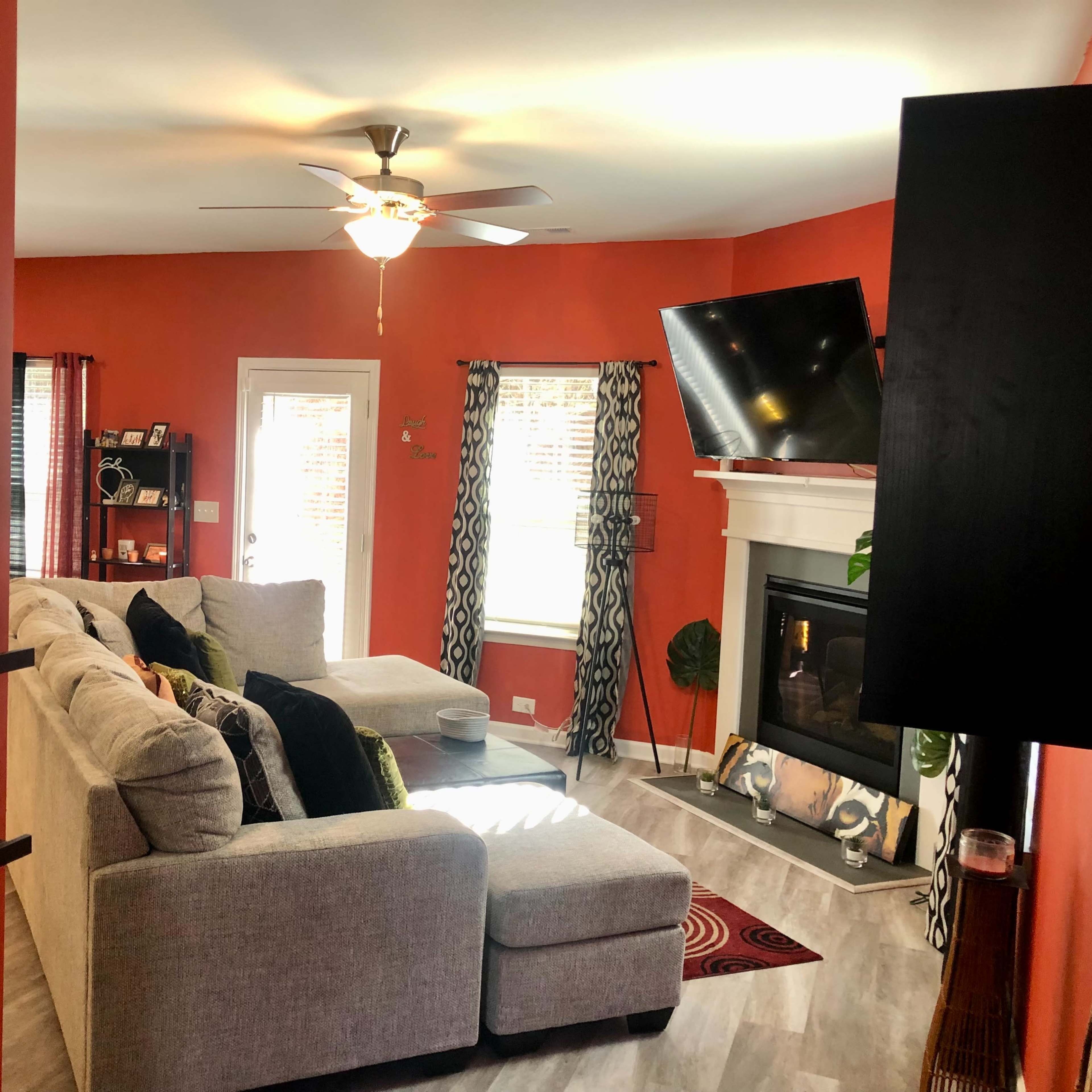 A cozy living room features a gray sectional sofa, a ceiling fan, and a television mounted above a fireplace, with orange walls and decorative accents.