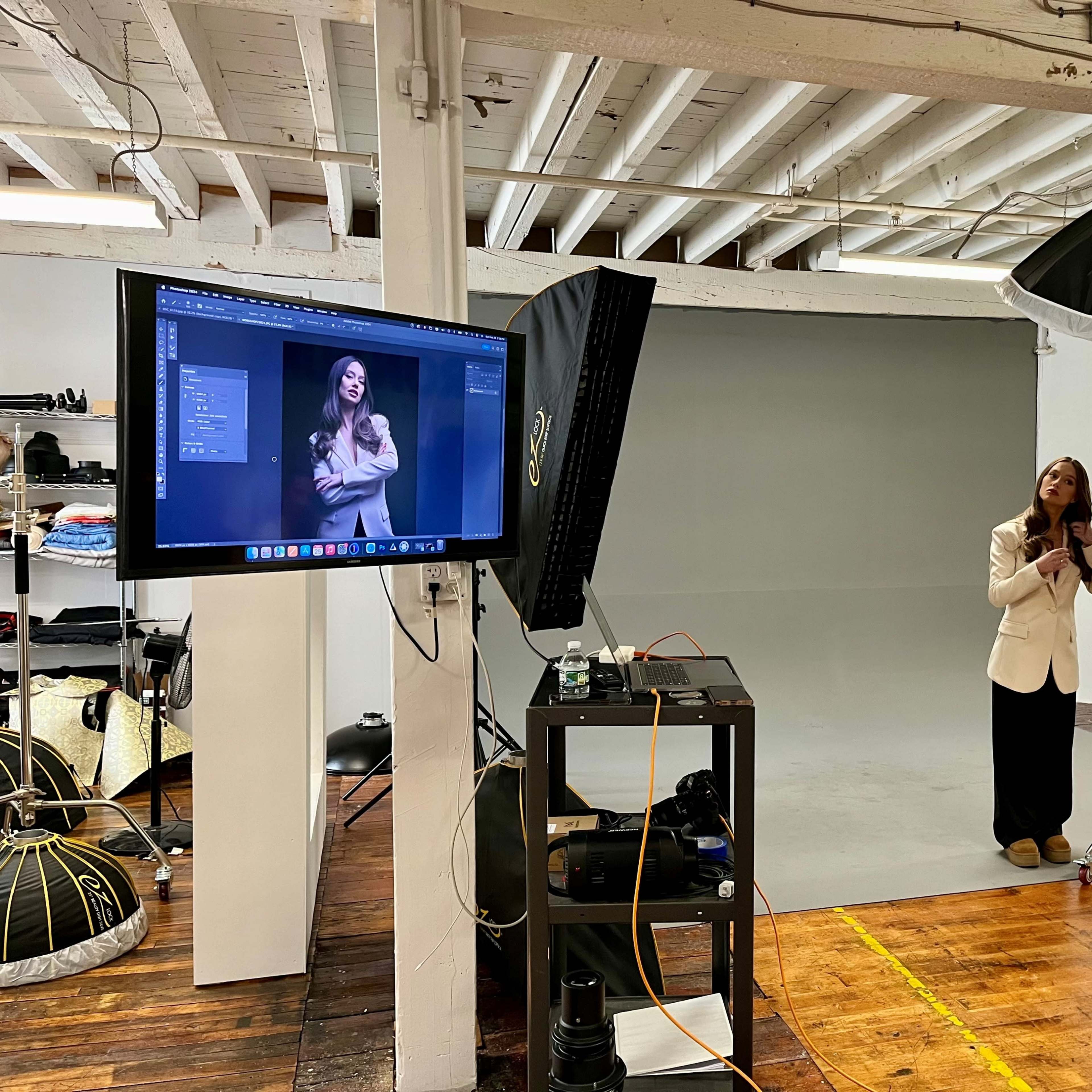 A woman in a white blazer poses in a photography studio with a gray backdrop while a monitor displays her image.