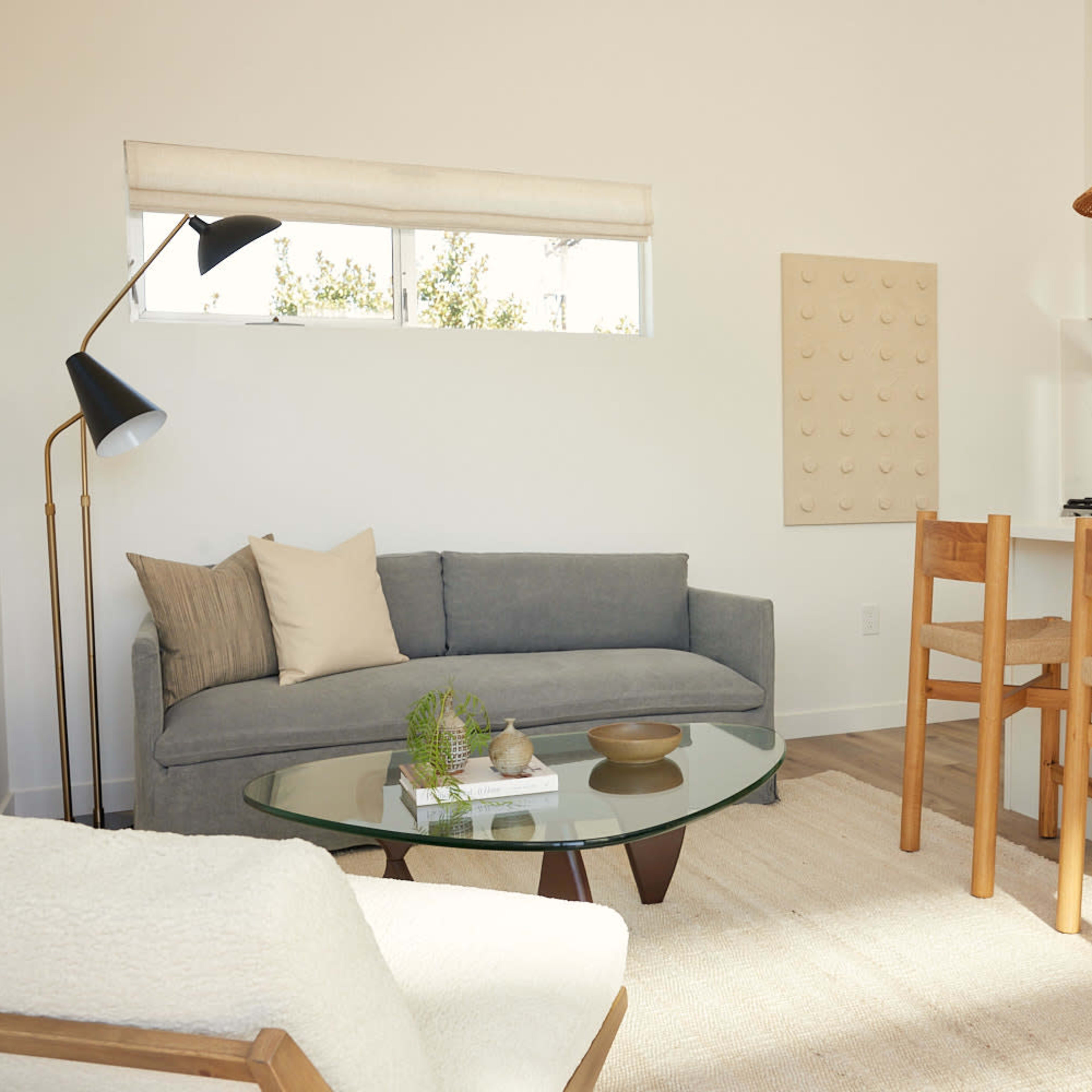 A modern living space features a gray sofa, a glass coffee table, a white dining counter with two wooden chairs, and natural light coming through the windows.