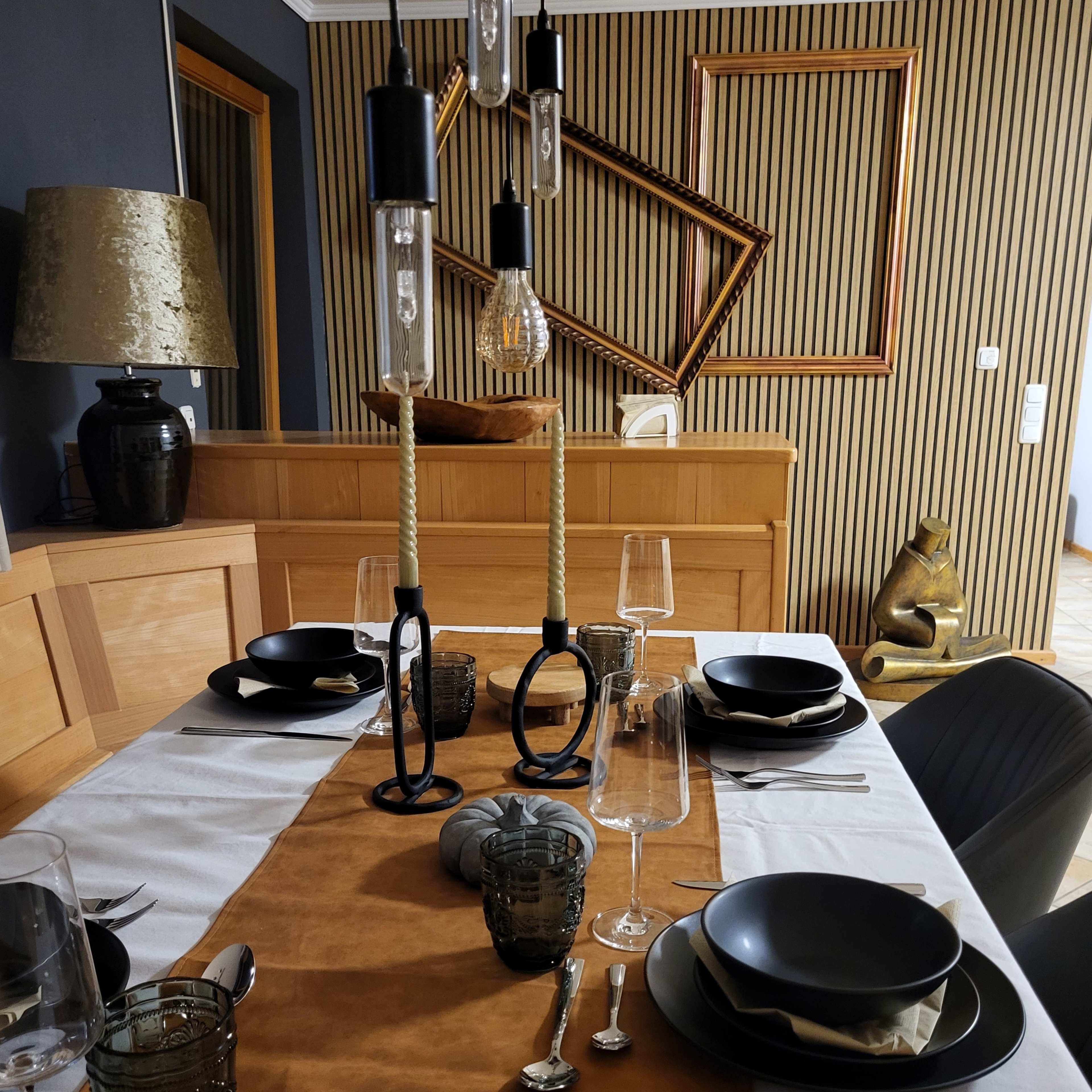 A dining table is set with black dishware and glassware, featuring a brown table runner, in a room with striped wallpaper and decorative frames.