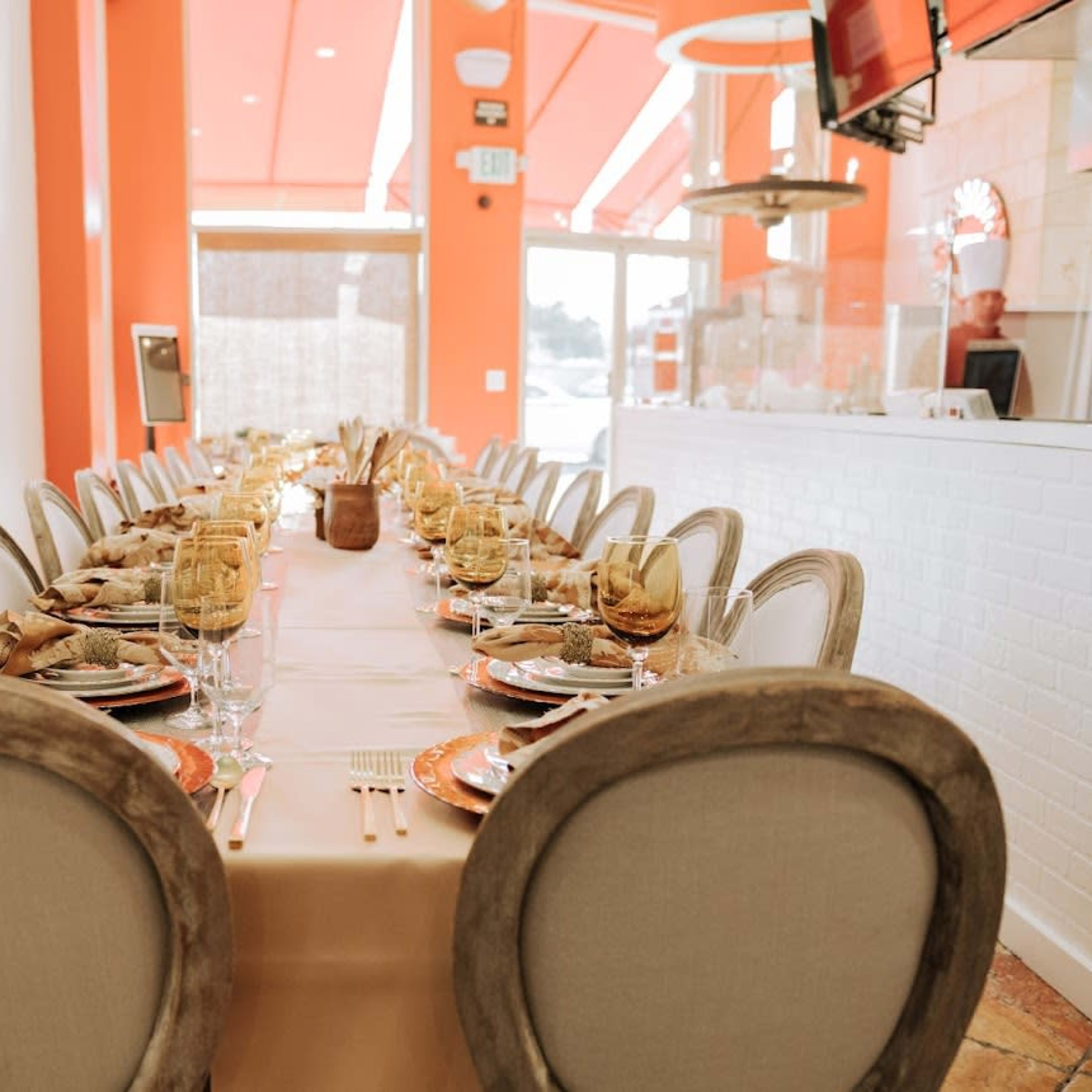 A long dining table is set with plates, glasses, and napkins under orange walls in a brightly lit restaurant.