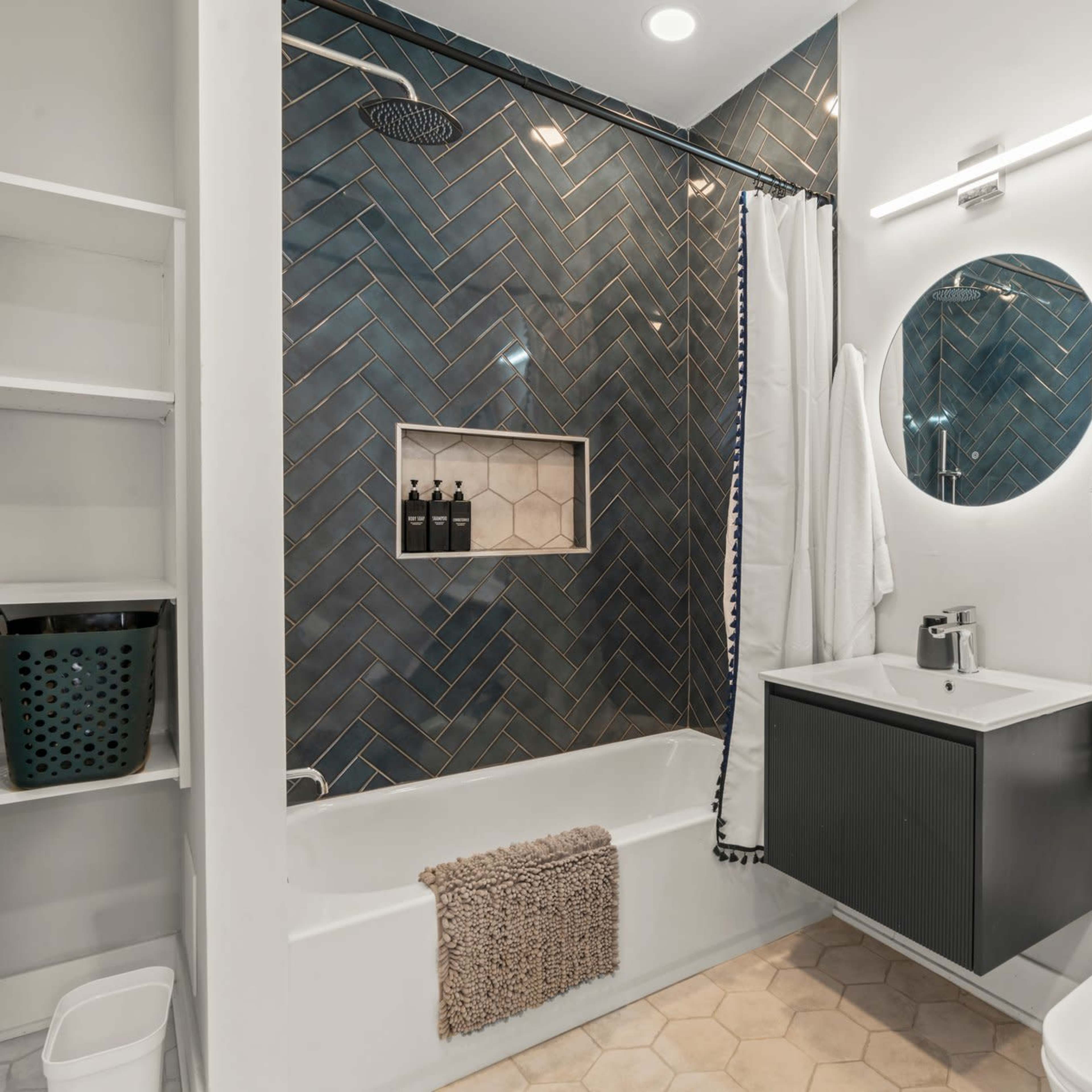 A modern bathroom featuring a shower-tub combination with a herringbone tile pattern, a circular mirror, and a compact sink with storage below.