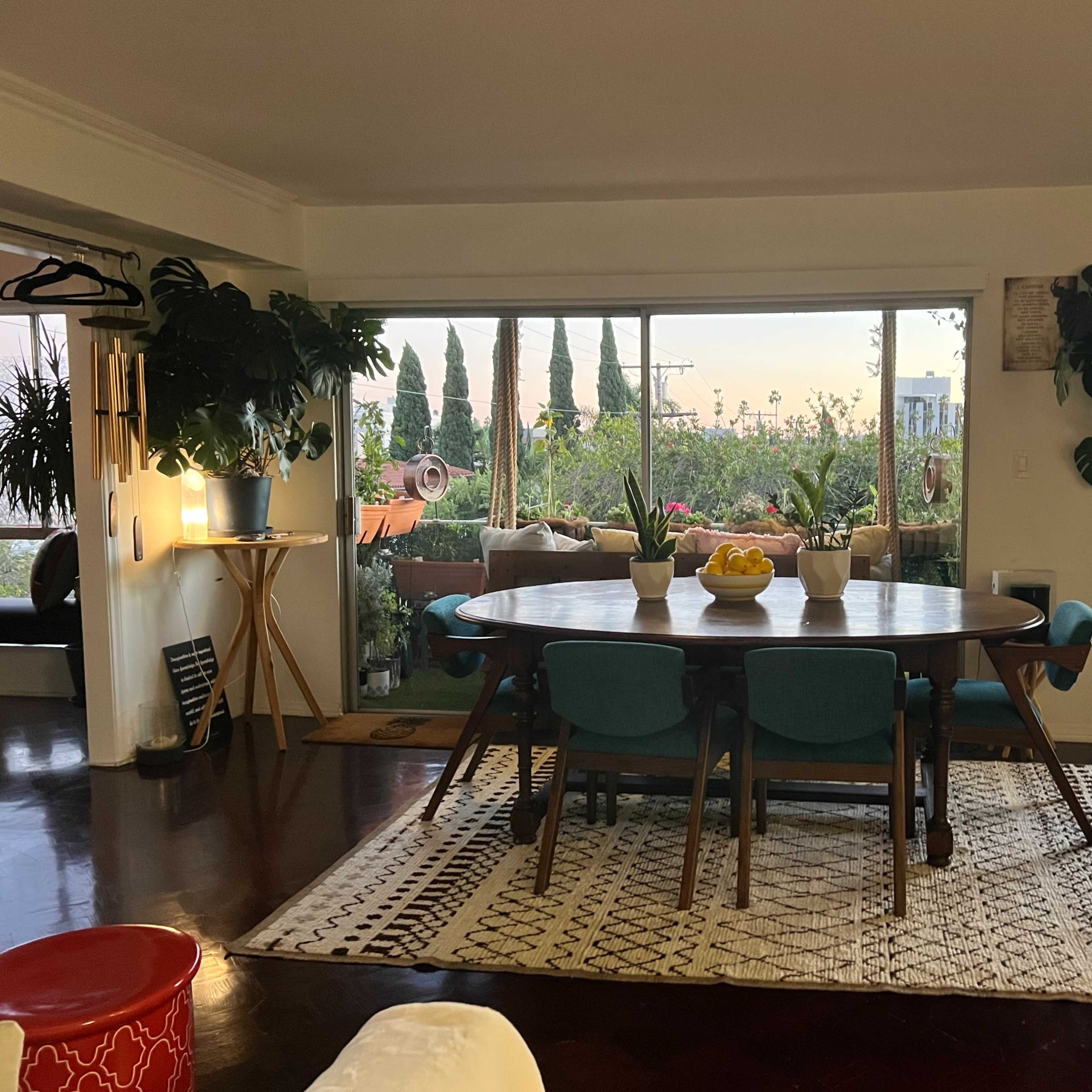 A spacious dining area features a large round table surrounded by teal chairs, with plants and large windows providing natural light.