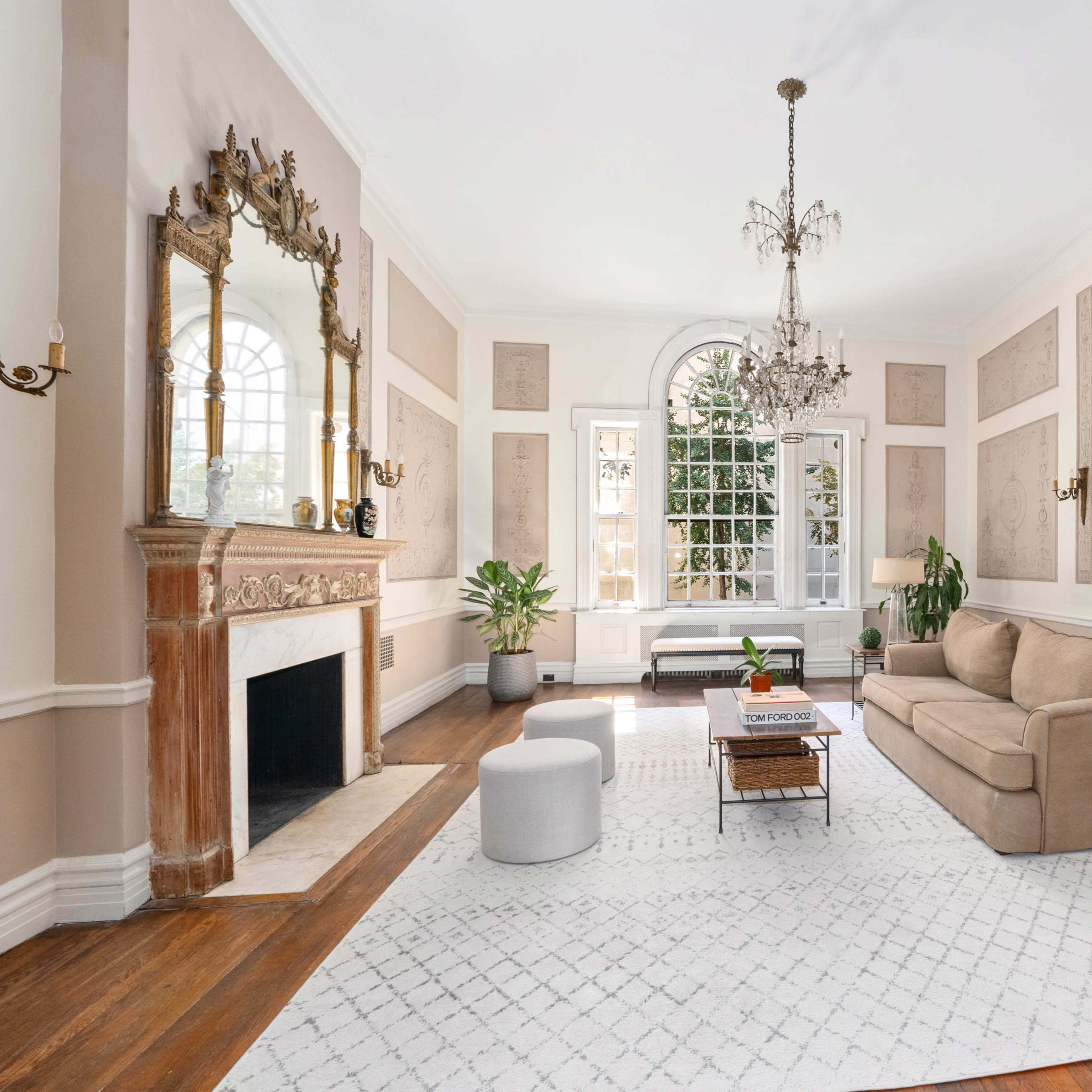 A spacious living room with a beige sofa, a patterned area rug, a marble fireplace, and large windows allowing natural light.