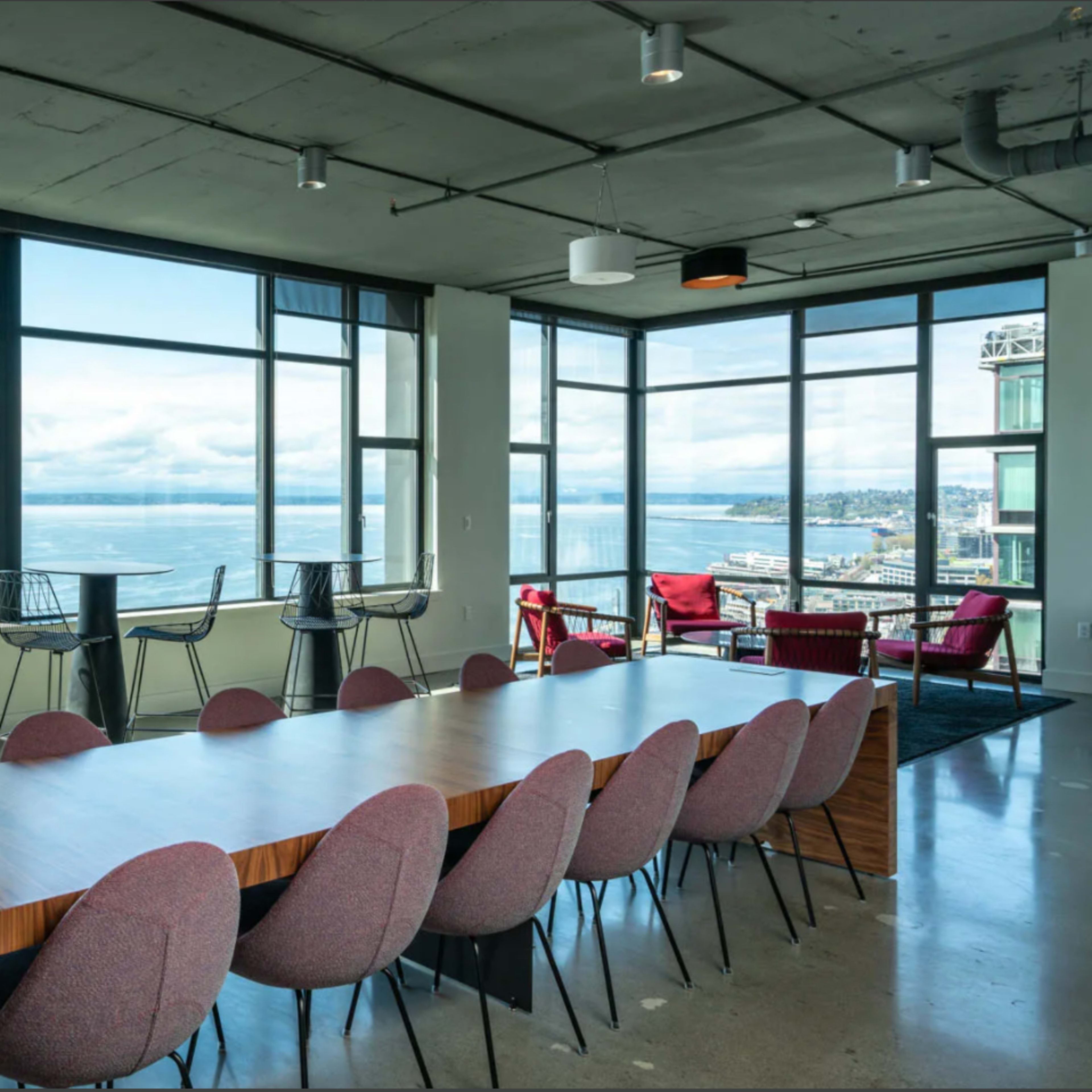 A modern conference room with a long wooden table and eight patterned chairs, featuring large windows that offer a view of the water and cloudy sky outside.