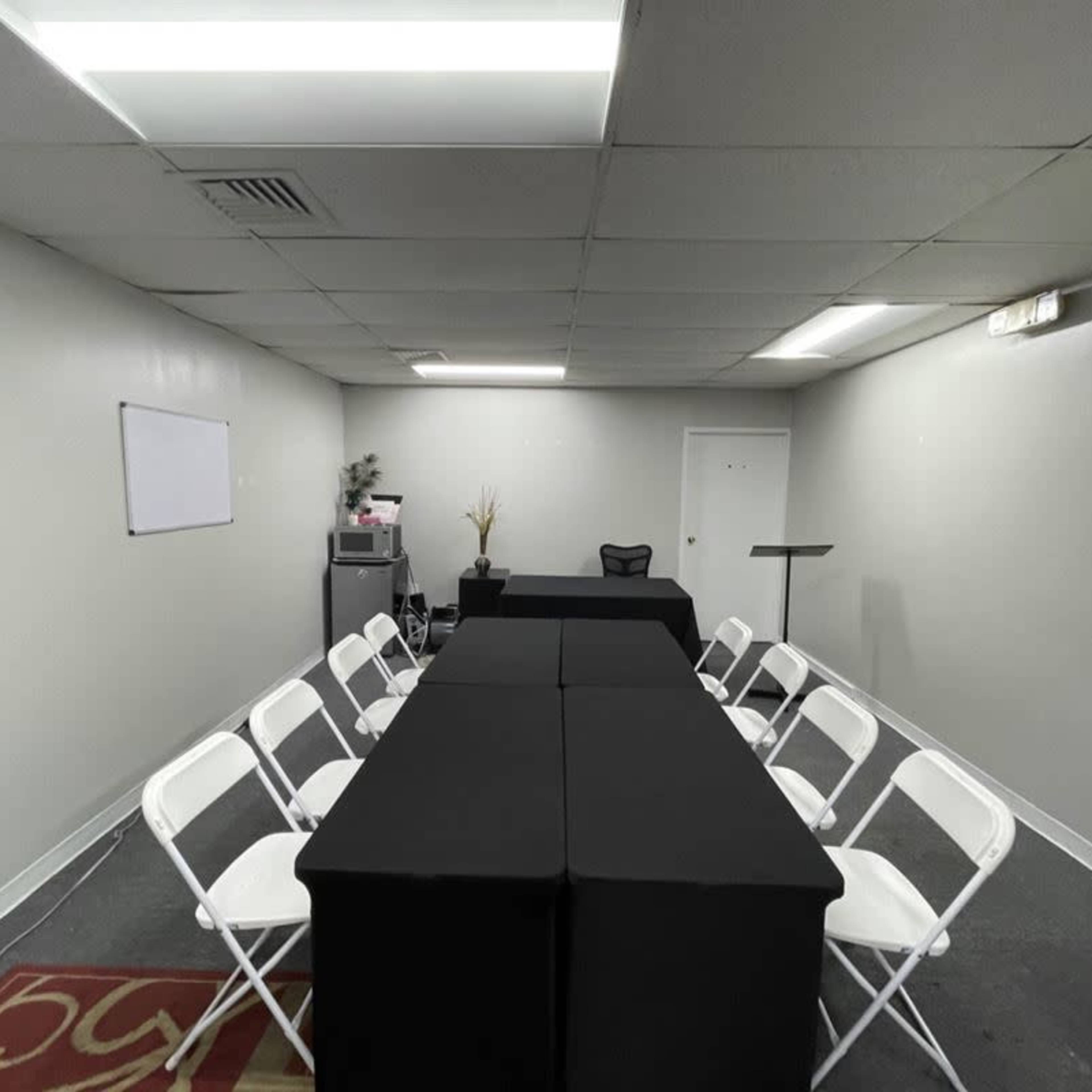 A meeting room arranged with two rectangular tables covered in black cloth, surrounded by white folding chairs, and featuring a dry-erase board and a small table in the corner.