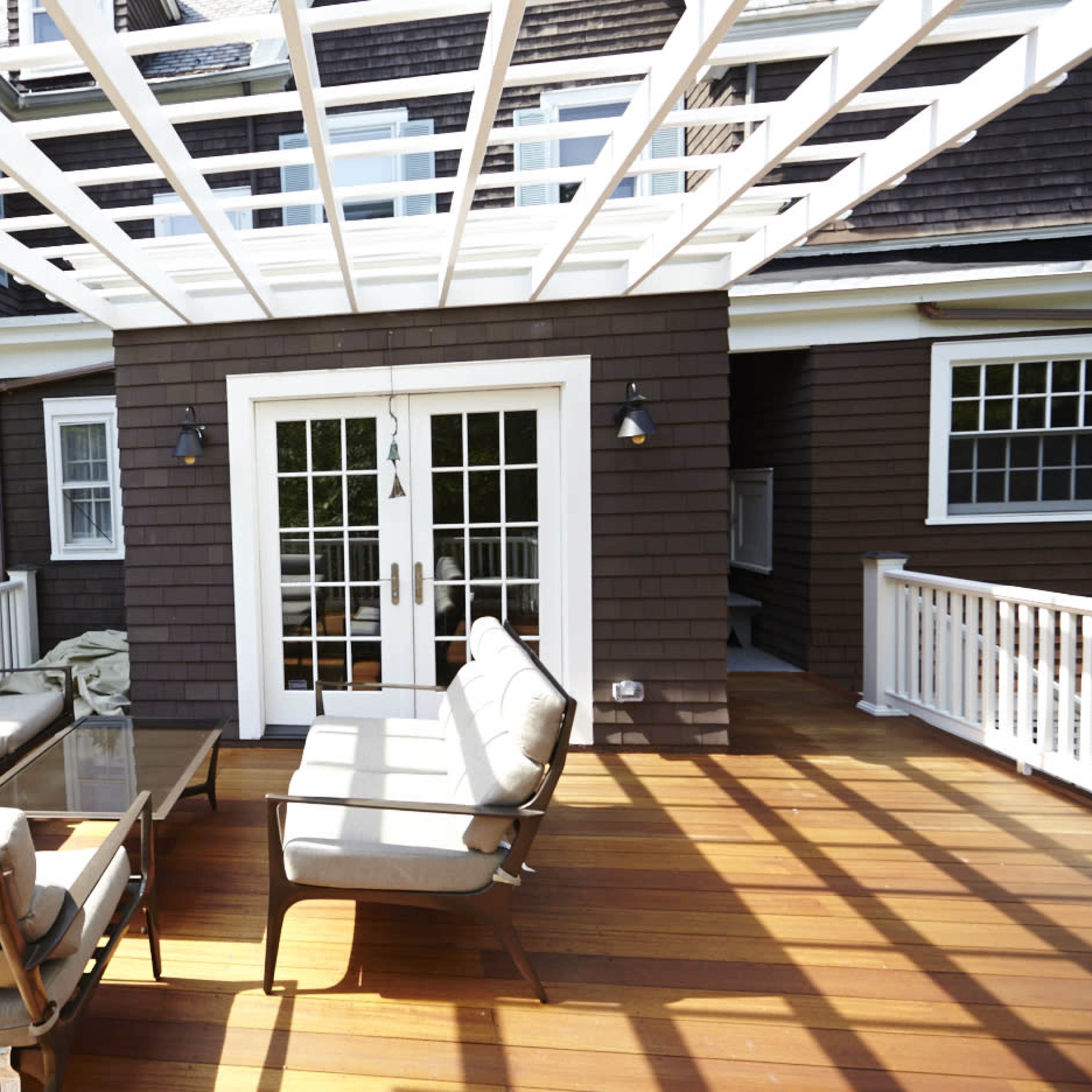 A wooden deck with a seating area that includes a sofa and glass table, covered by a white pergola.