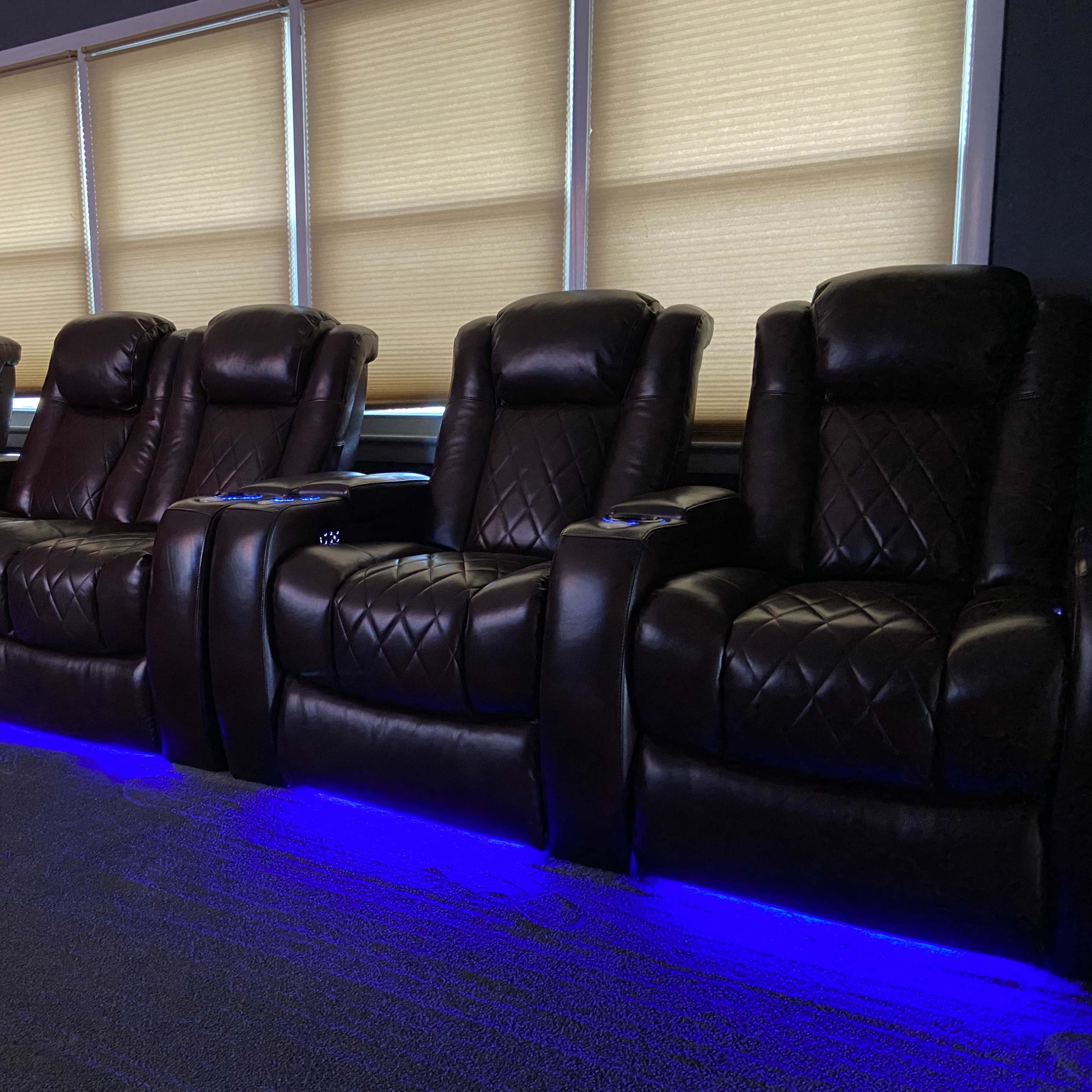 A row of leather recliners with blue LED lighting is arranged in front of large windows with shades.