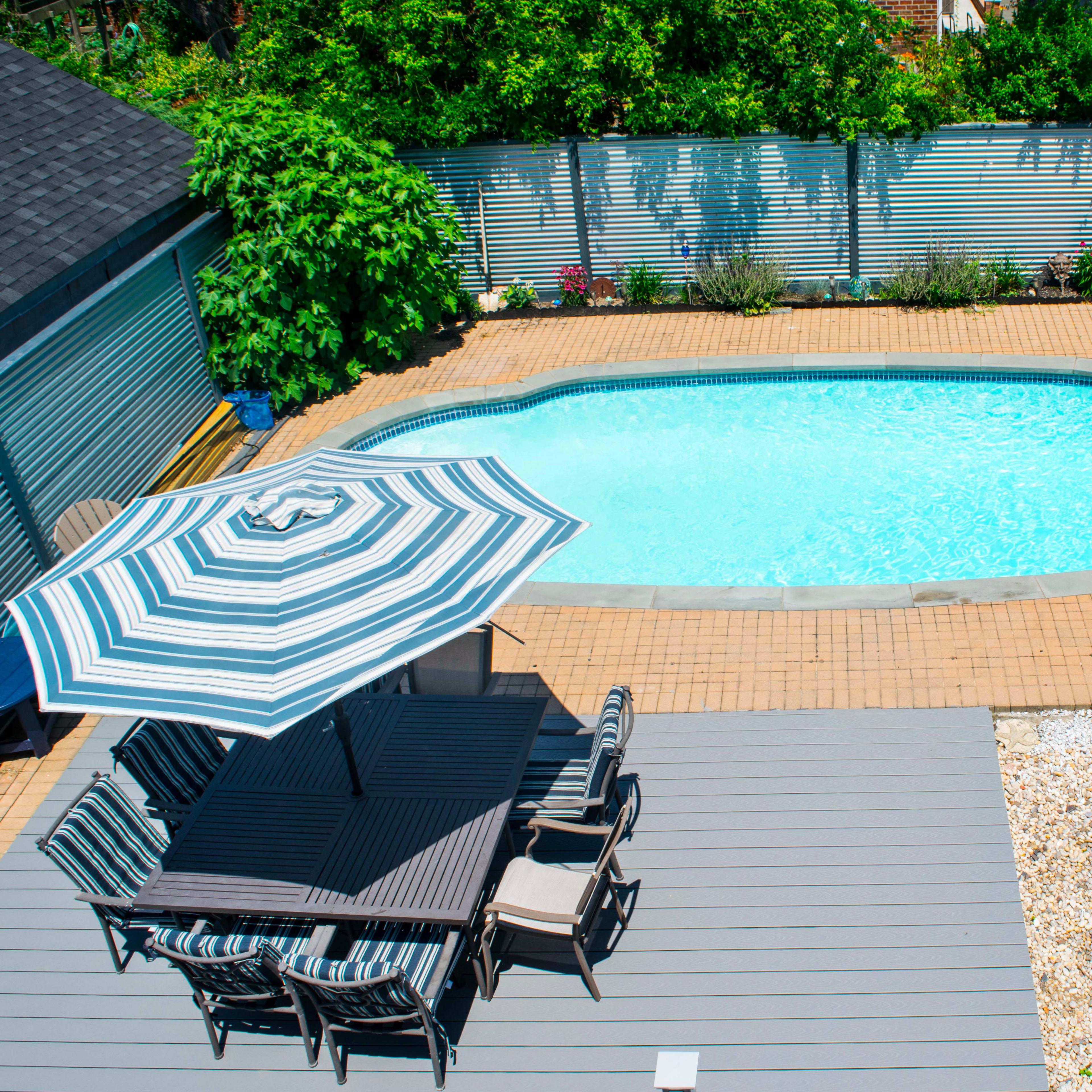 A rectangular swimming pool surrounded by a patio with lounge chairs and a dining area shaded by a striped umbrella.