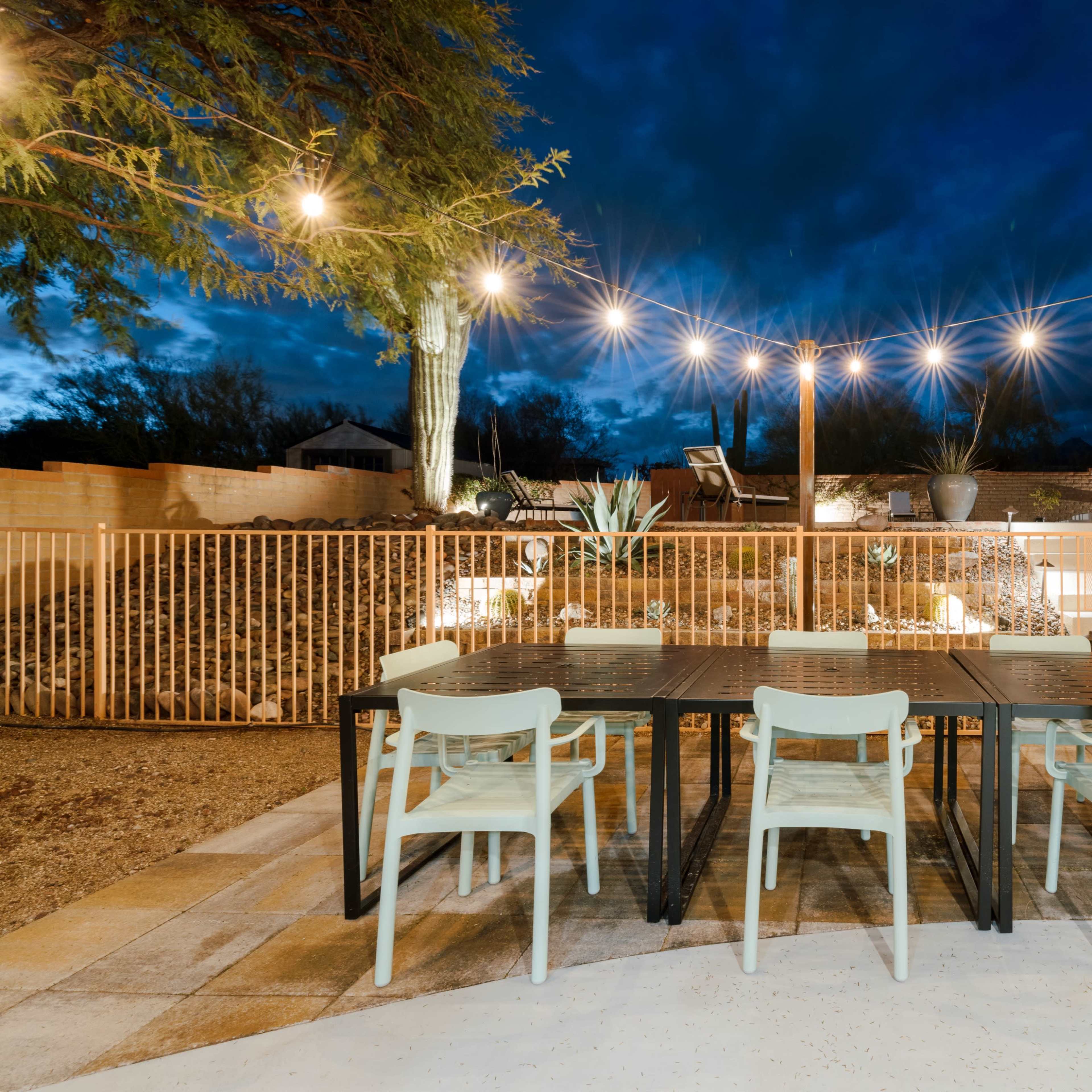 A dining area with a large table and chairs is illuminated by string lights against a nighttime backdrop of clouds and desert landscaping.