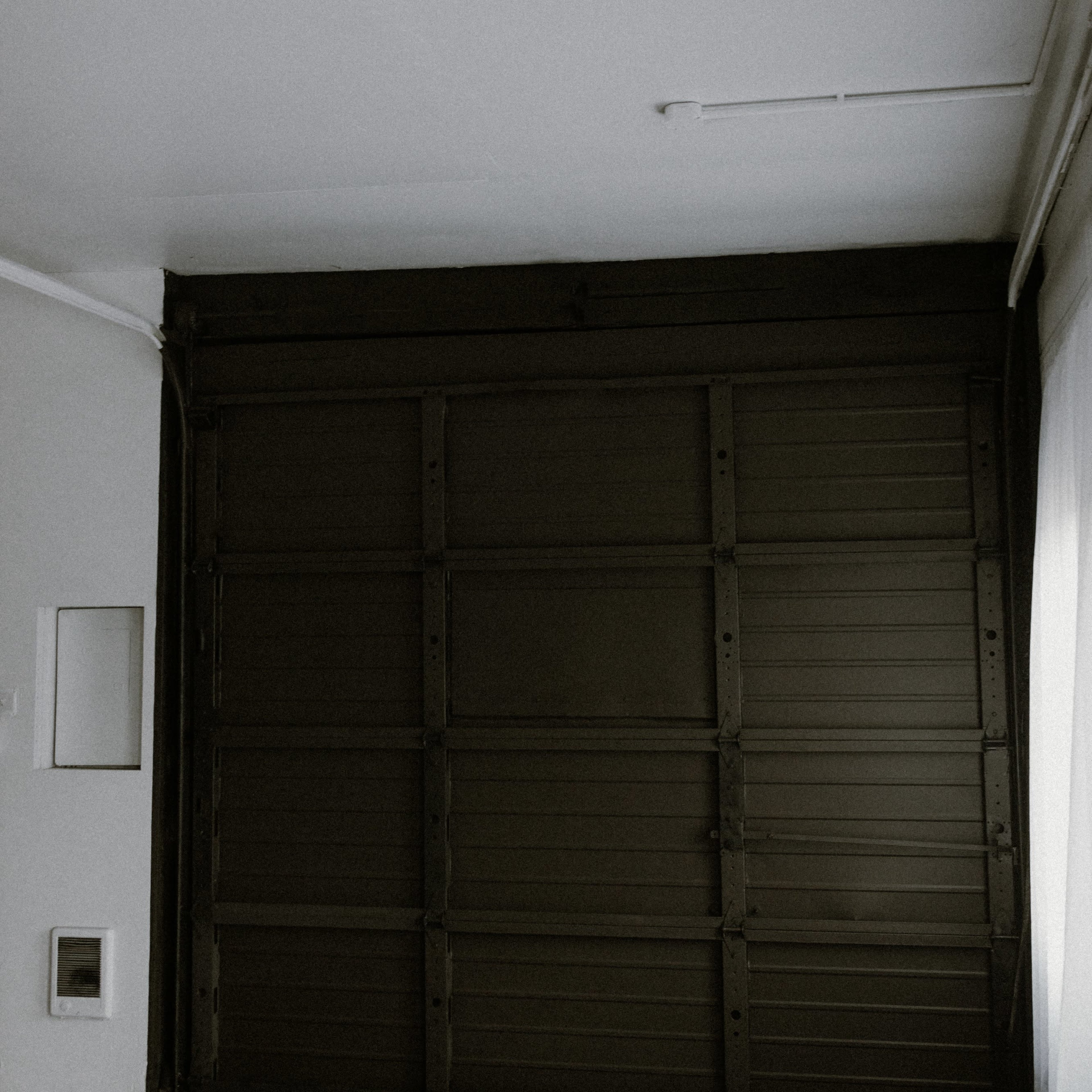 A minimalistic interior space with a large closed metal garage door and a white wall with a small square opening.