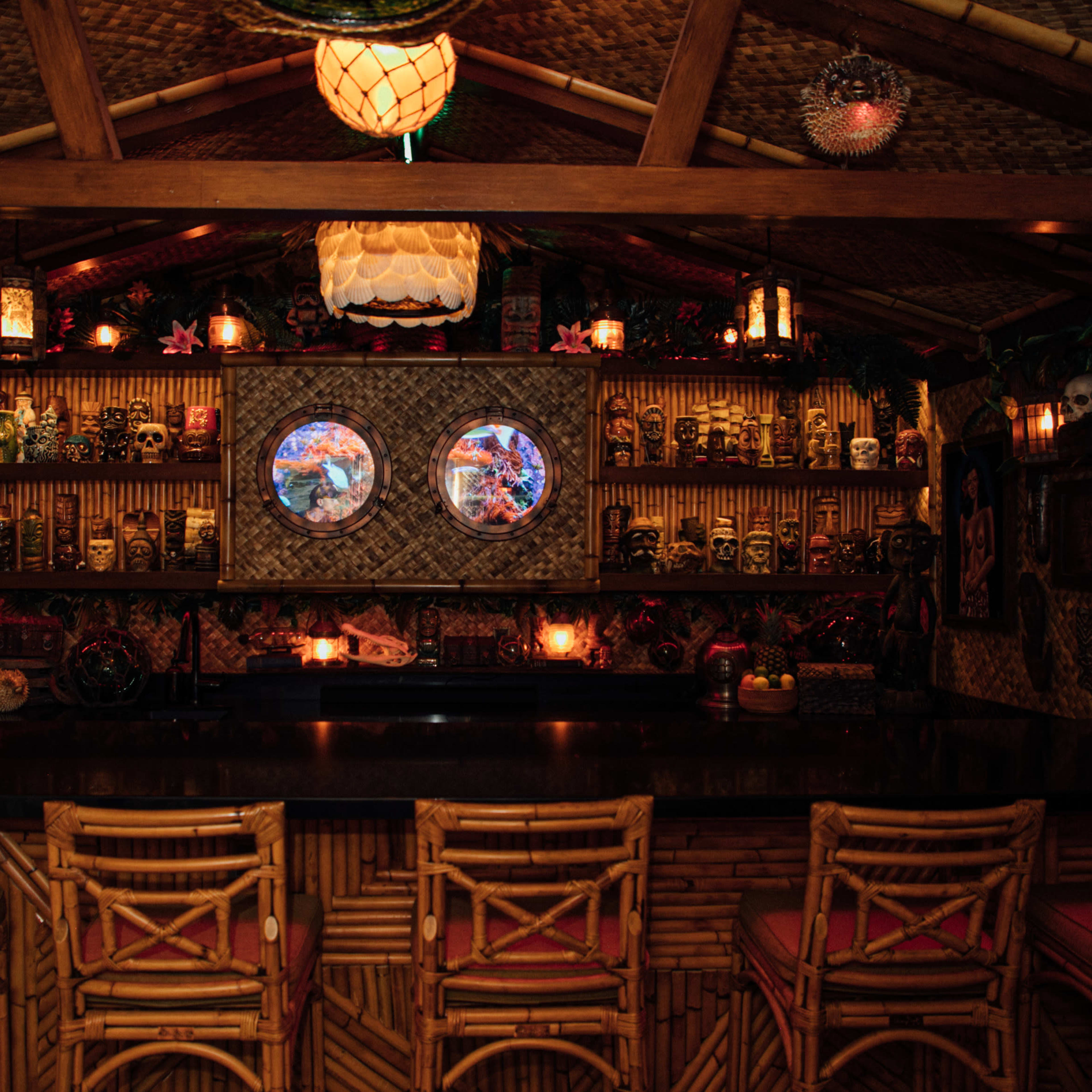 A tiki-themed bar with a bamboo structure, decorated shelves lined with various masks and skulls, and circular windows illuminated by colorful lights.