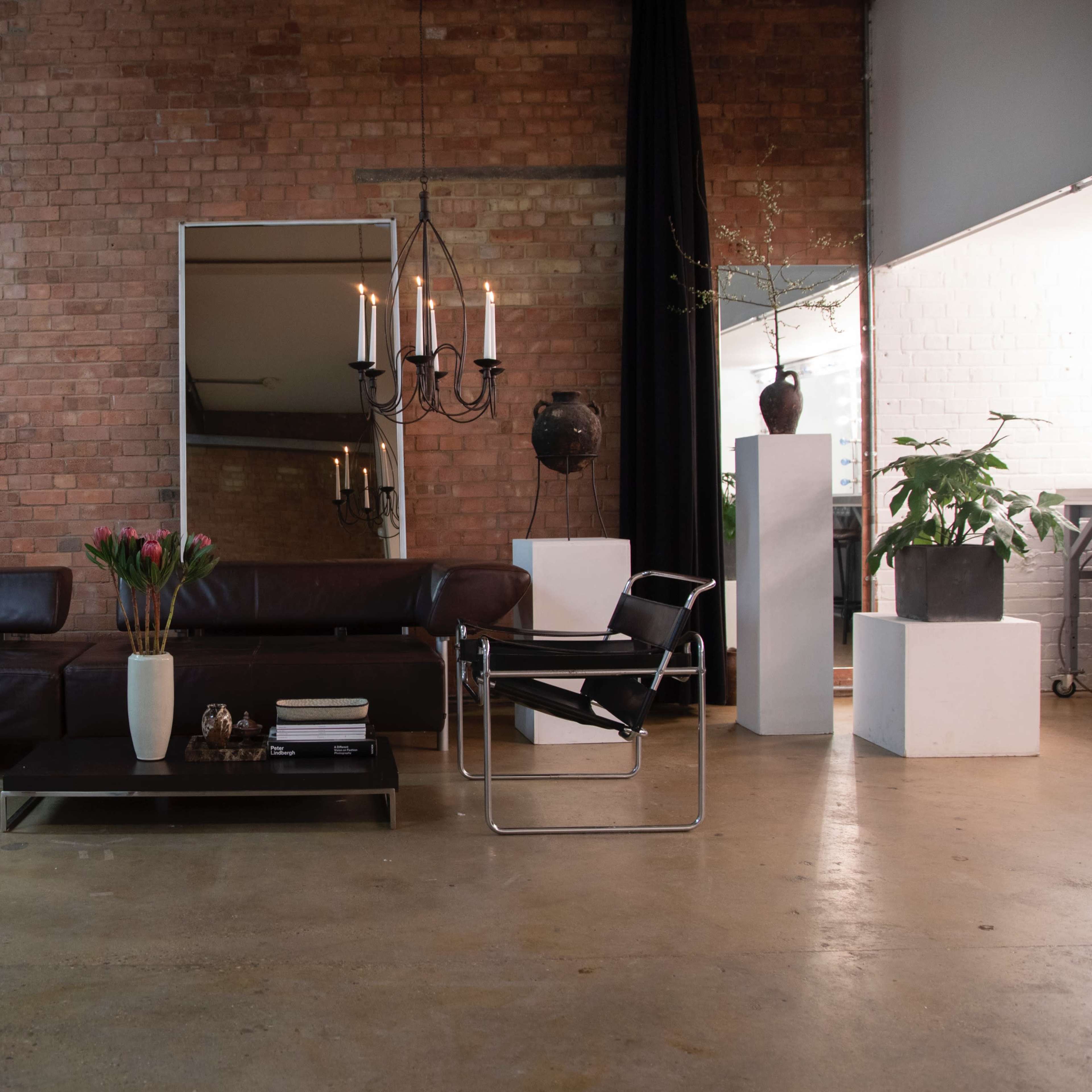 A modern living space featuring a dark leather sofa, a glass coffee table with decorative items, a large mirror, and plants against a brick wall.