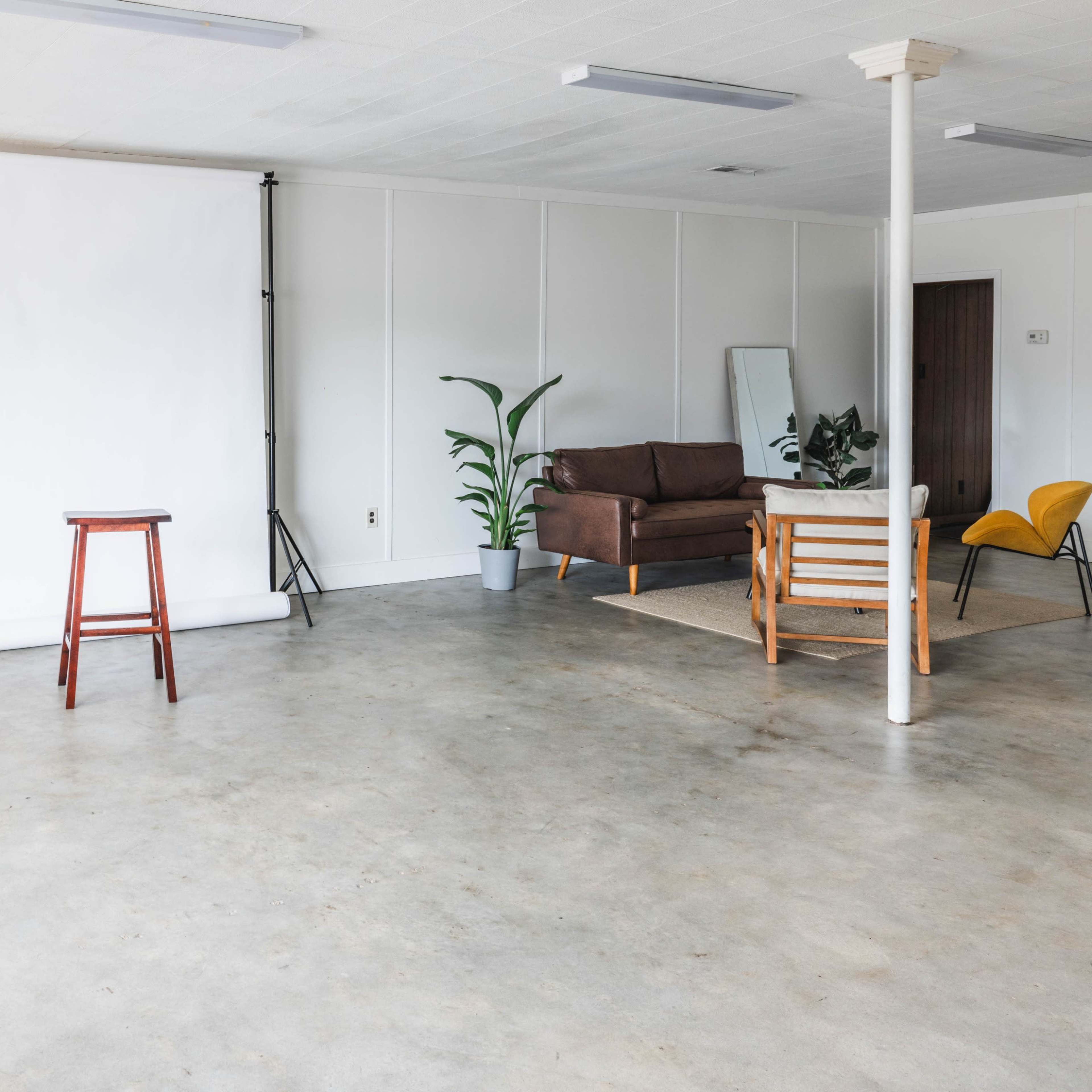 An empty room with a white backdrop, a wooden stool, a brown sofa, a yellow chair, and a potted plant.