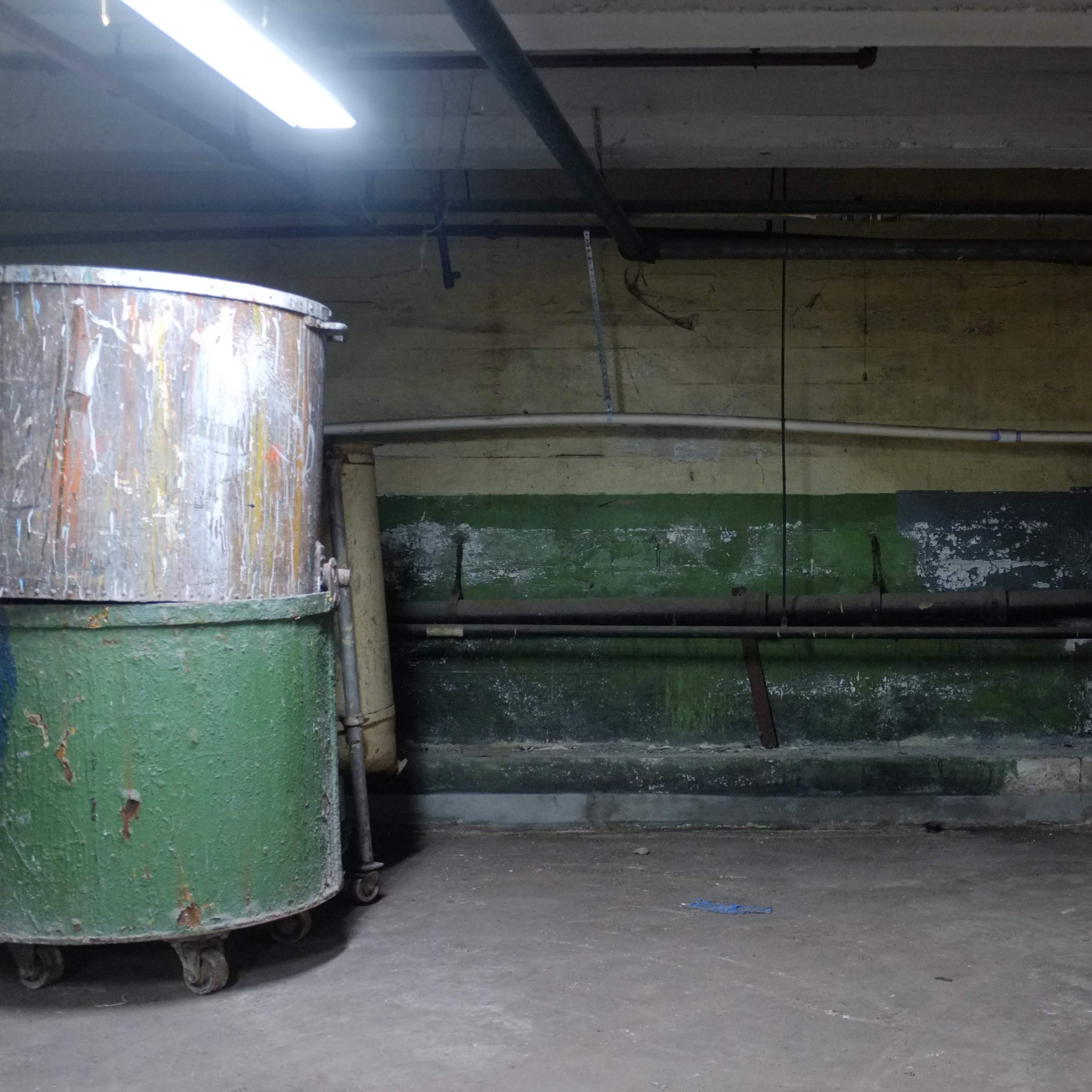 A dimly lit basement with a large metal drum and wooden crates stacked against the wall.