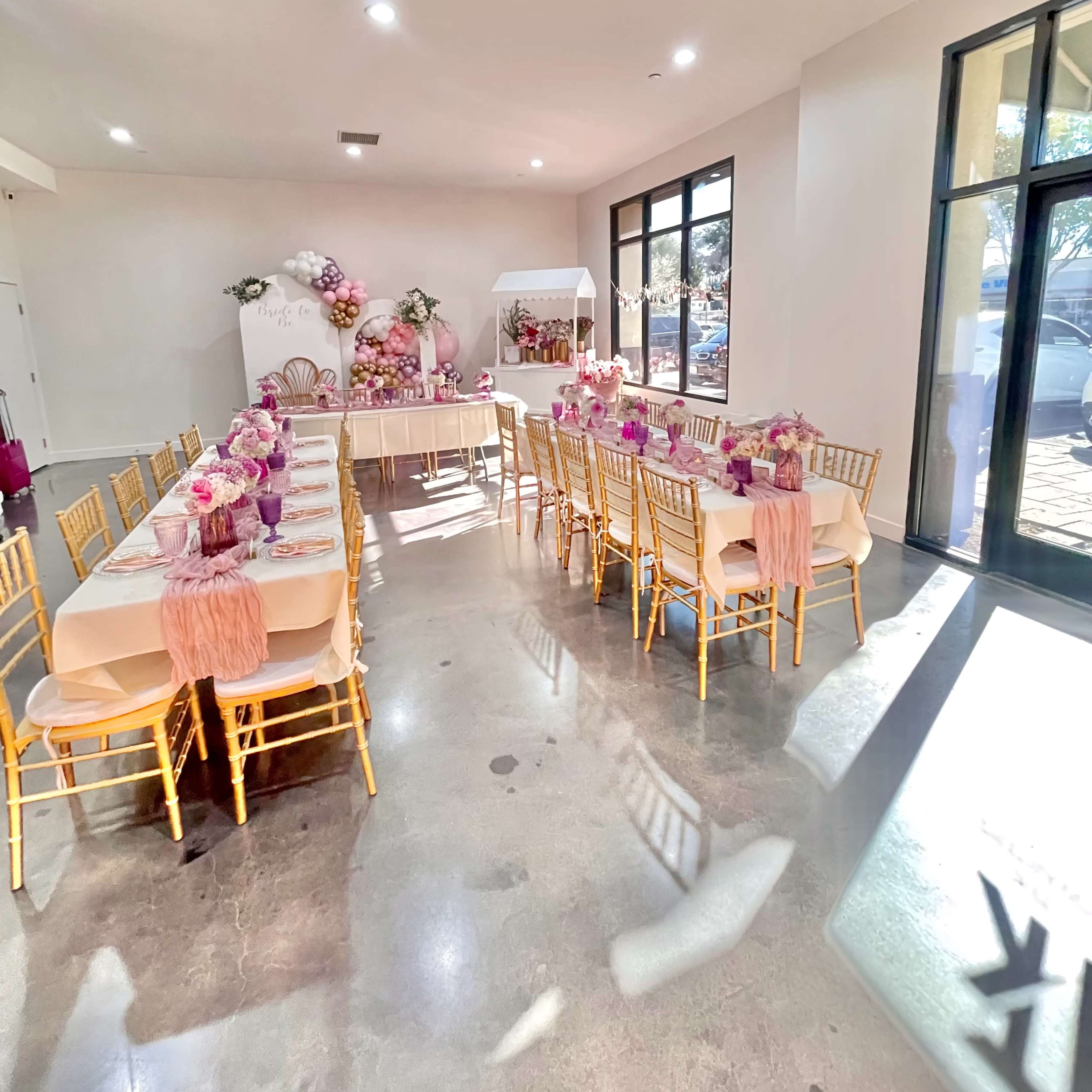 A decorated event space with tables set for a gathering, featuring pink floral arrangements and gold chairs.