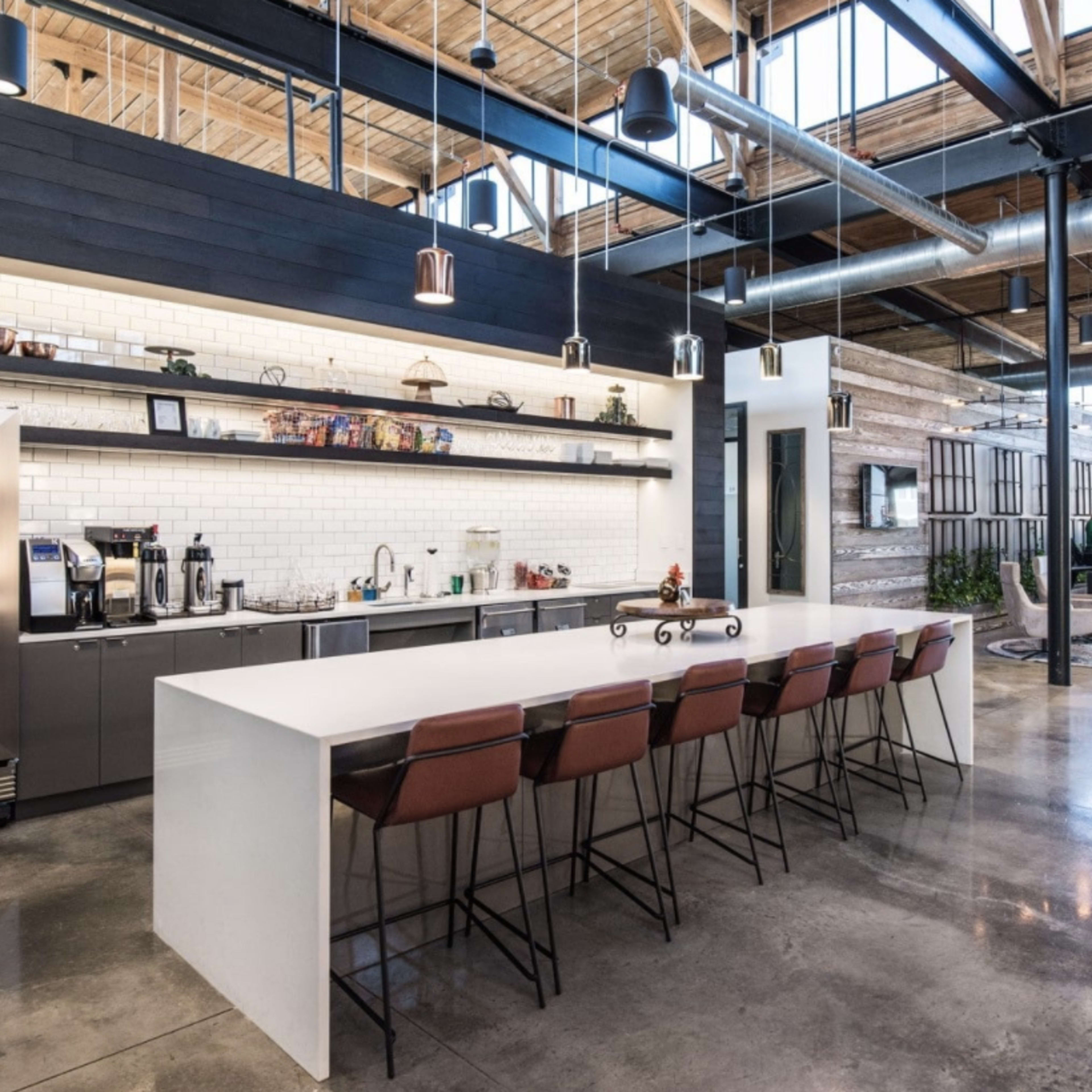 A modern kitchen area with a long white countertop, high-backed chairs, and stainless steel appliances against a backdrop of exposed wooden beams and polished concrete floors.