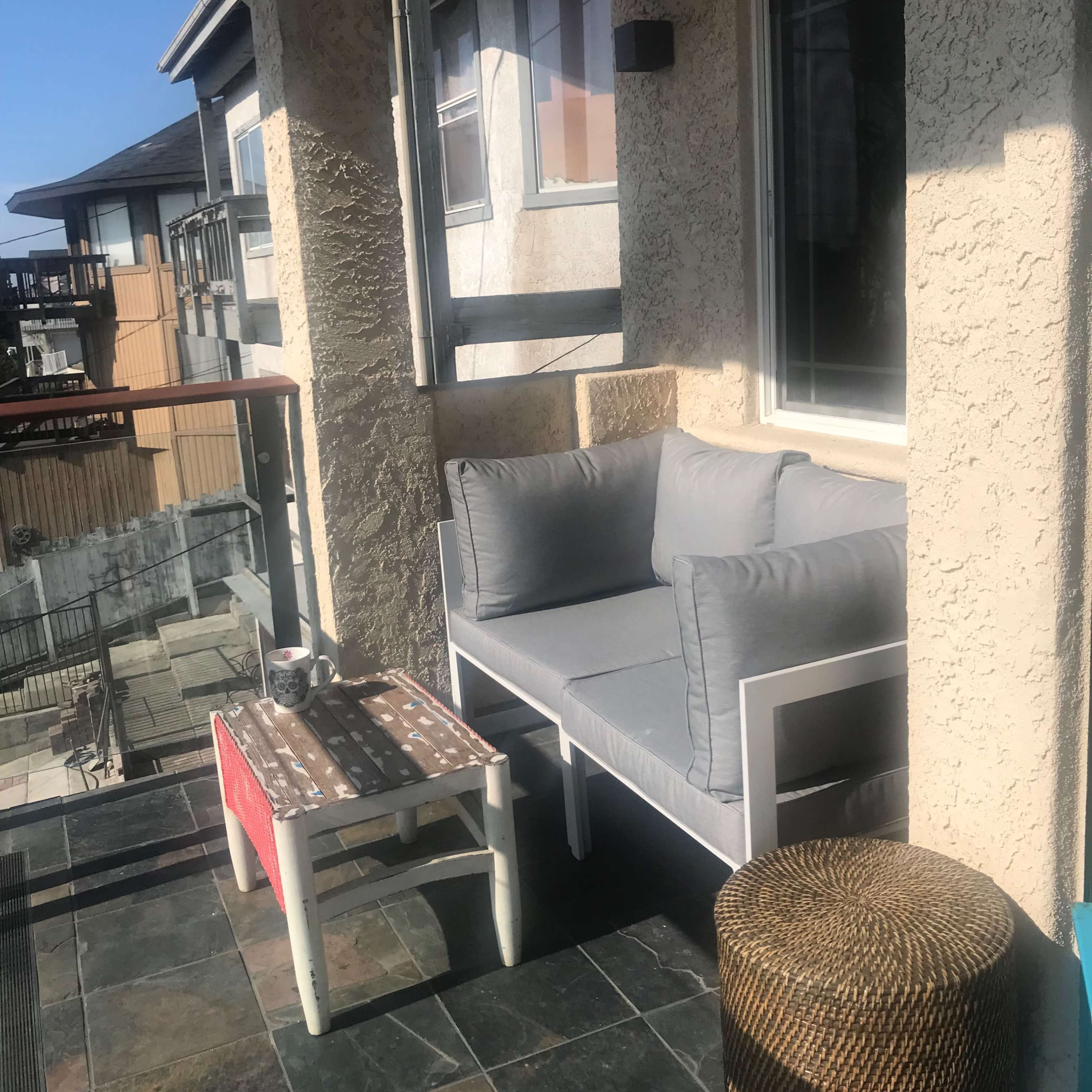 A small balcony features a gray couch, a low table with a decorative cloth, and a wicker stool, surrounded by sunny outdoor views.