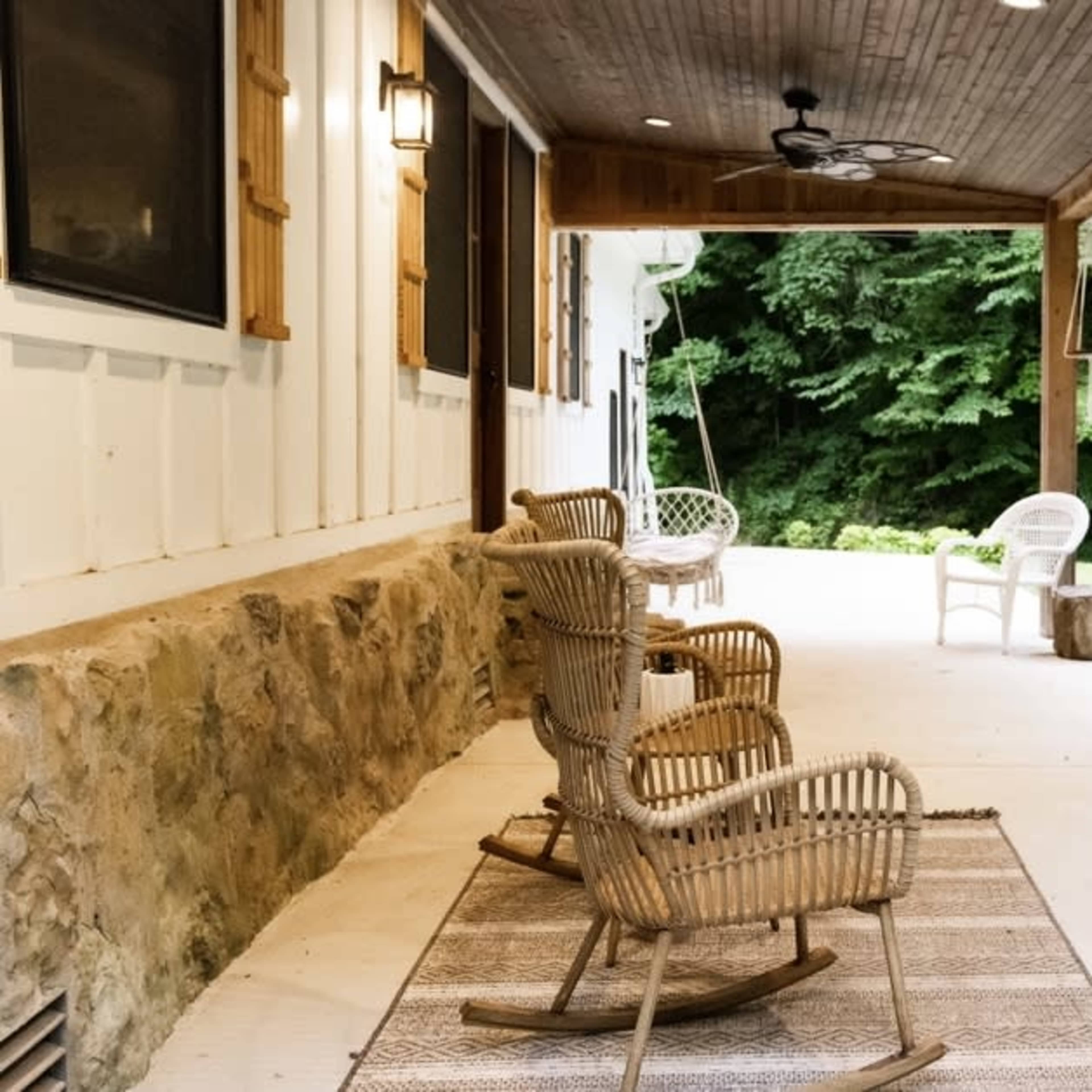 A covered porch featuring wicker rocking chairs and a swing, surrounded by green foliage.