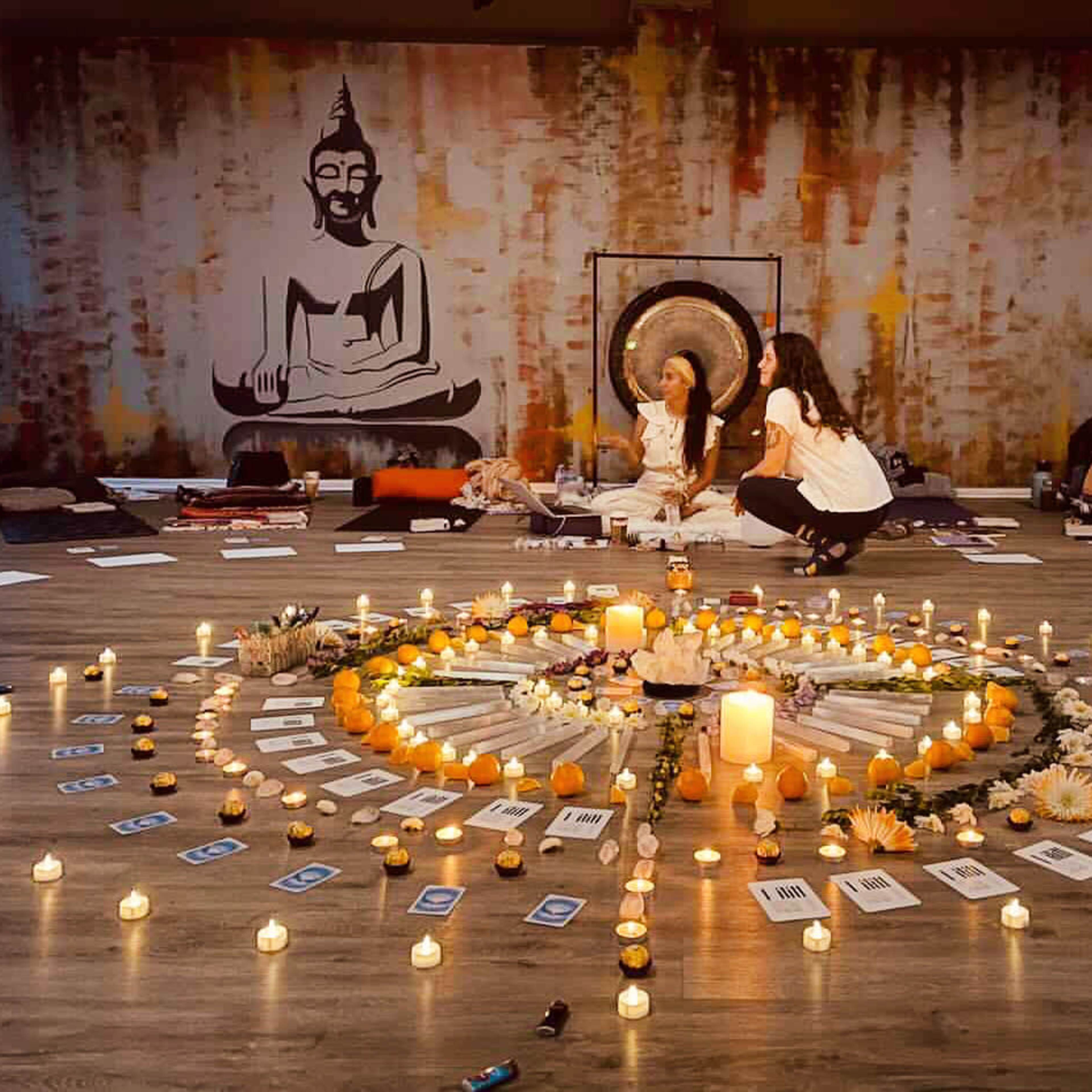 Two women sitting in a circular arrangement of candles, flowers, and cards in a dimly lit space decorated with a Buddha mural.