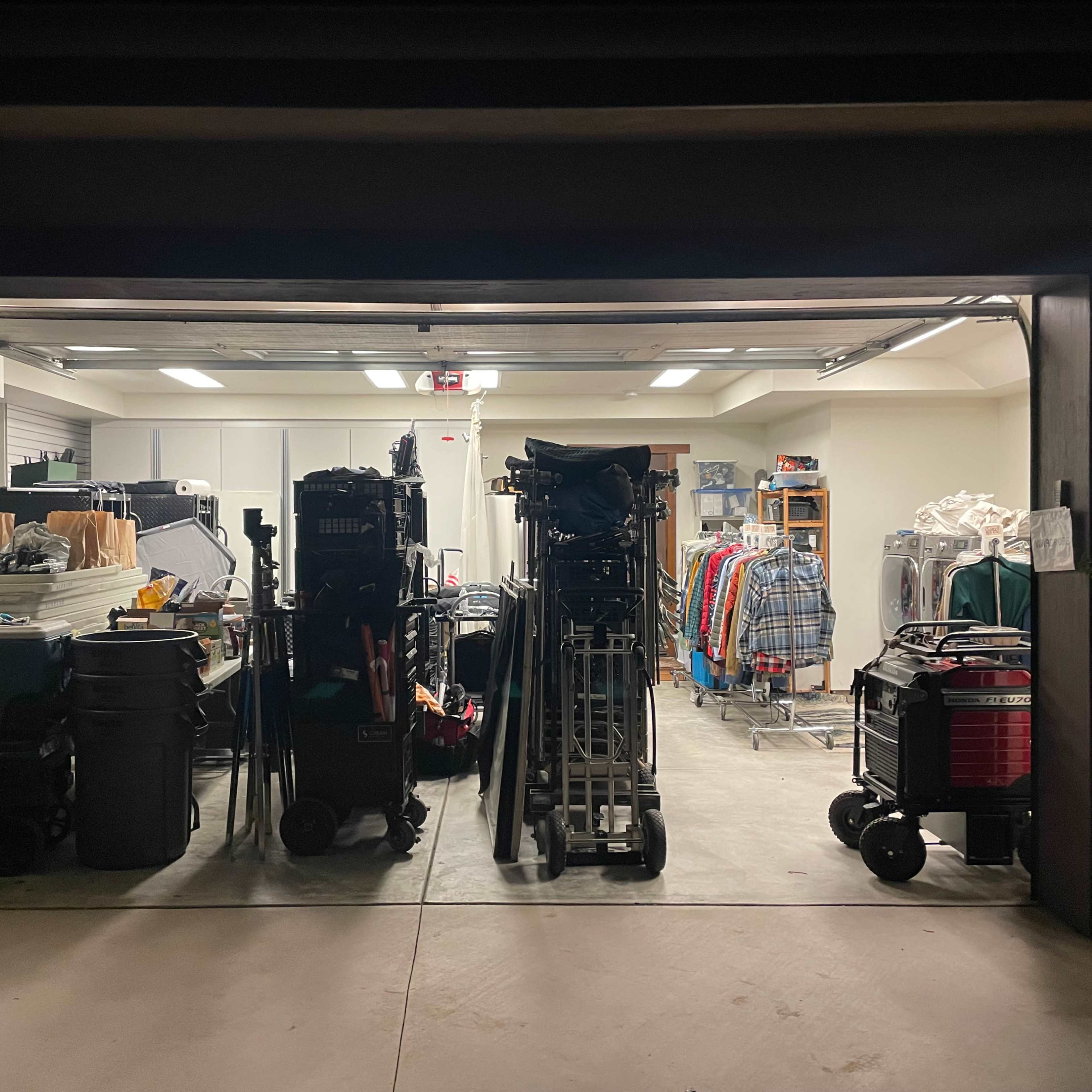 A dimly lit garage displays various storage items, including clothing racks, carts, and containers, lined against the walls.