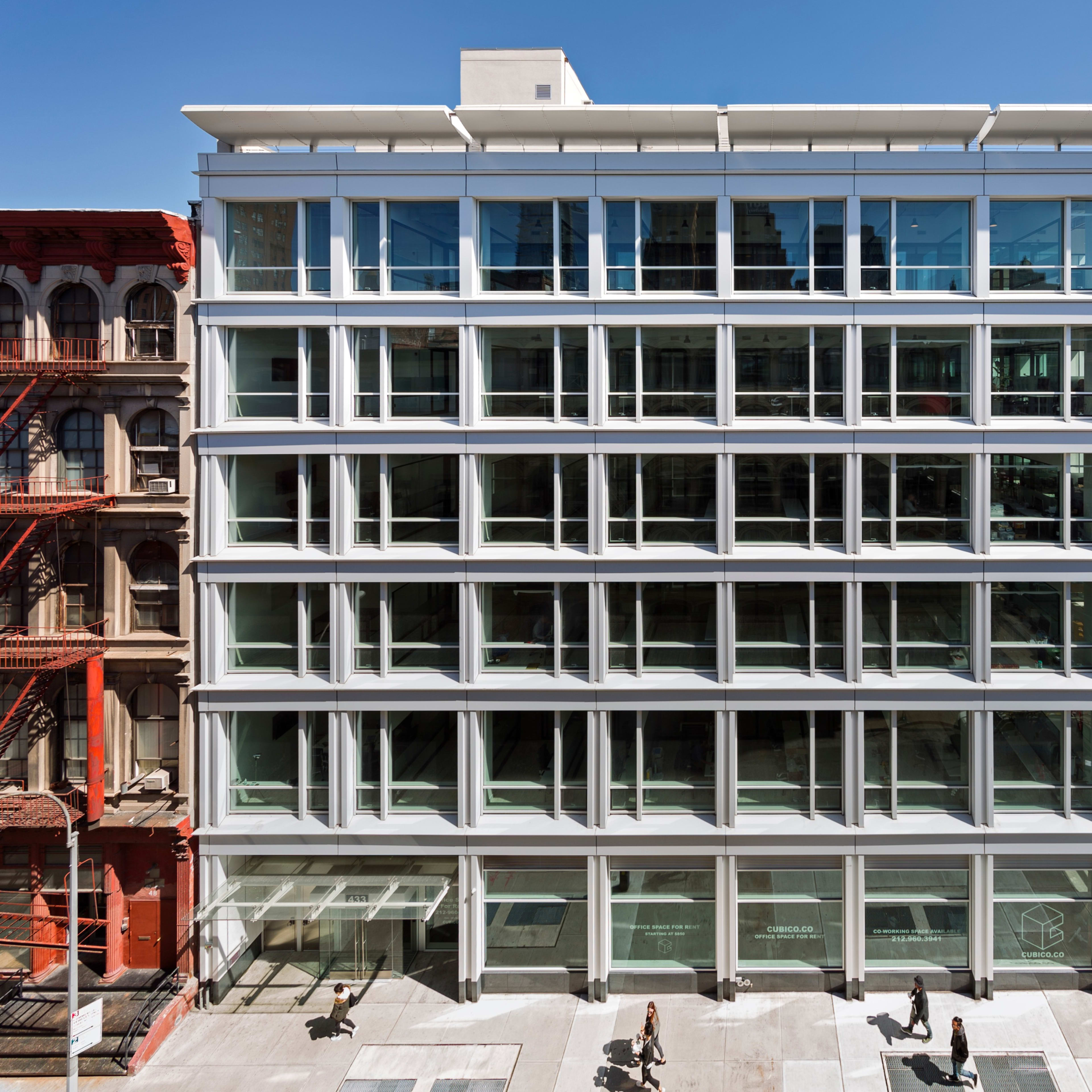 A modern, multi-story glass building features a grid of large windows facing a city street, with pedestrians walking by on the ground level.