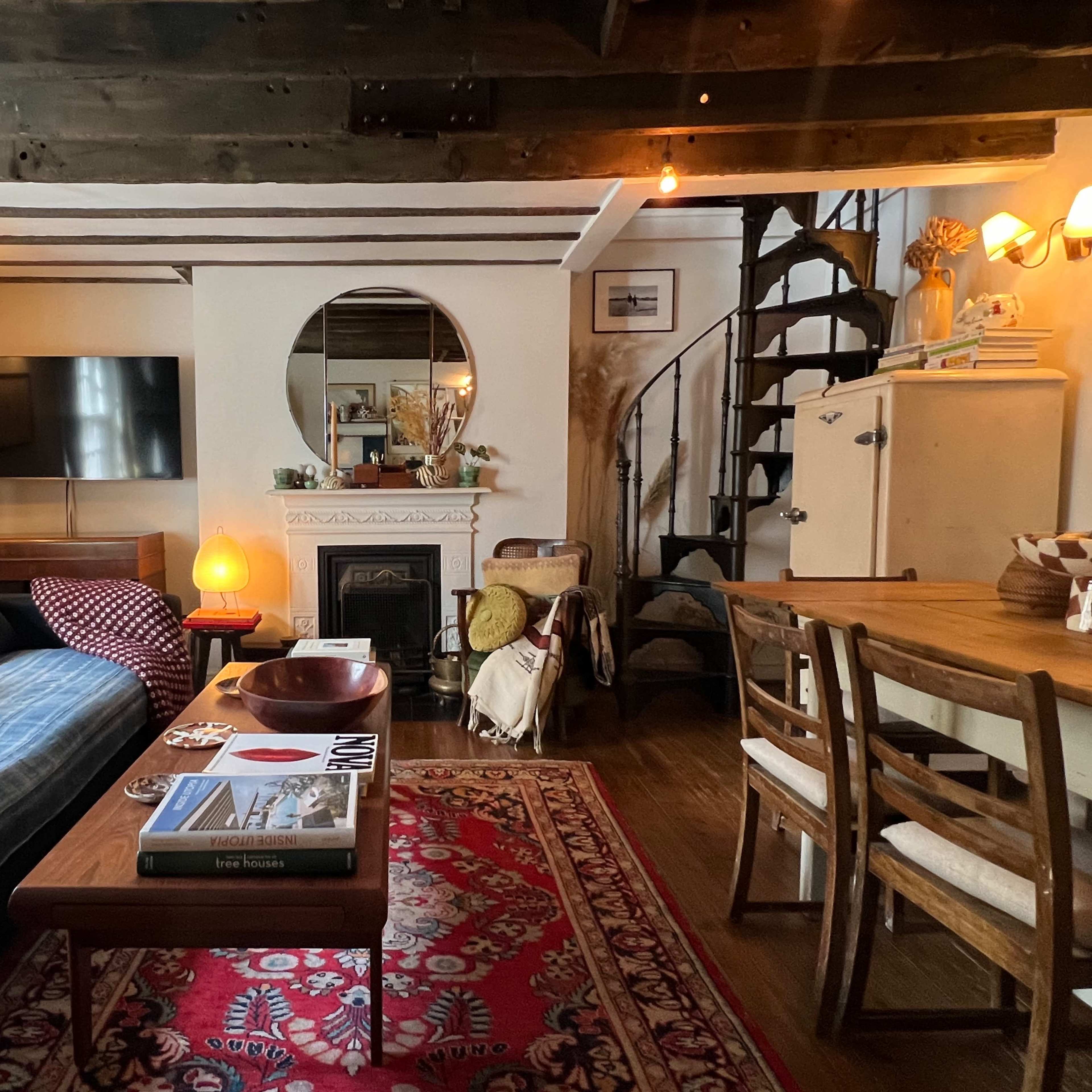 A cozy living room with a fireplace, a large mirror, a TV mounted on the wall, a staircase in the corner, and a wooden dining table surrounded by chairs.