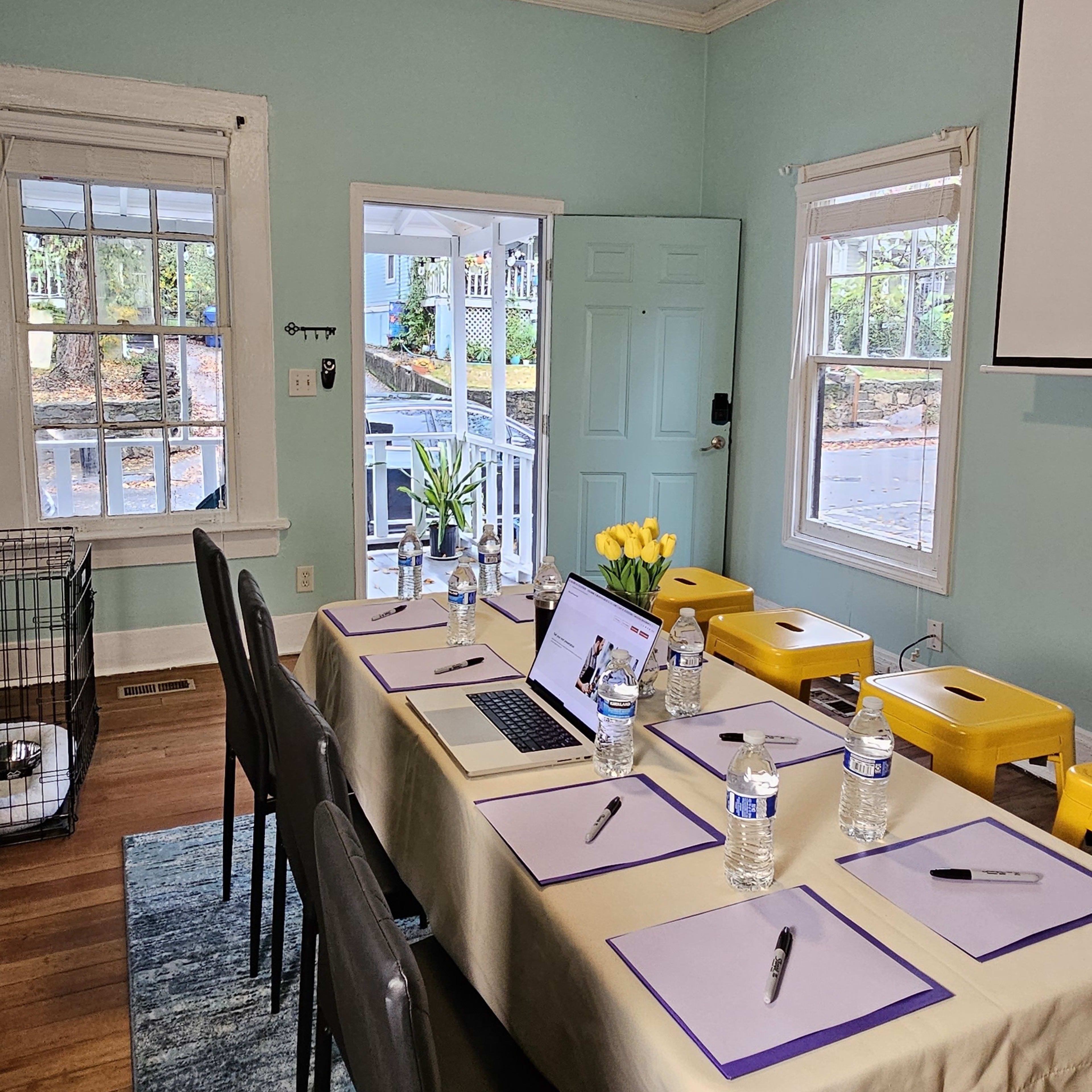 A meeting room is set up with a long table covered in papers, water bottles, and a laptop, flanked by chairs and a projector screen.
