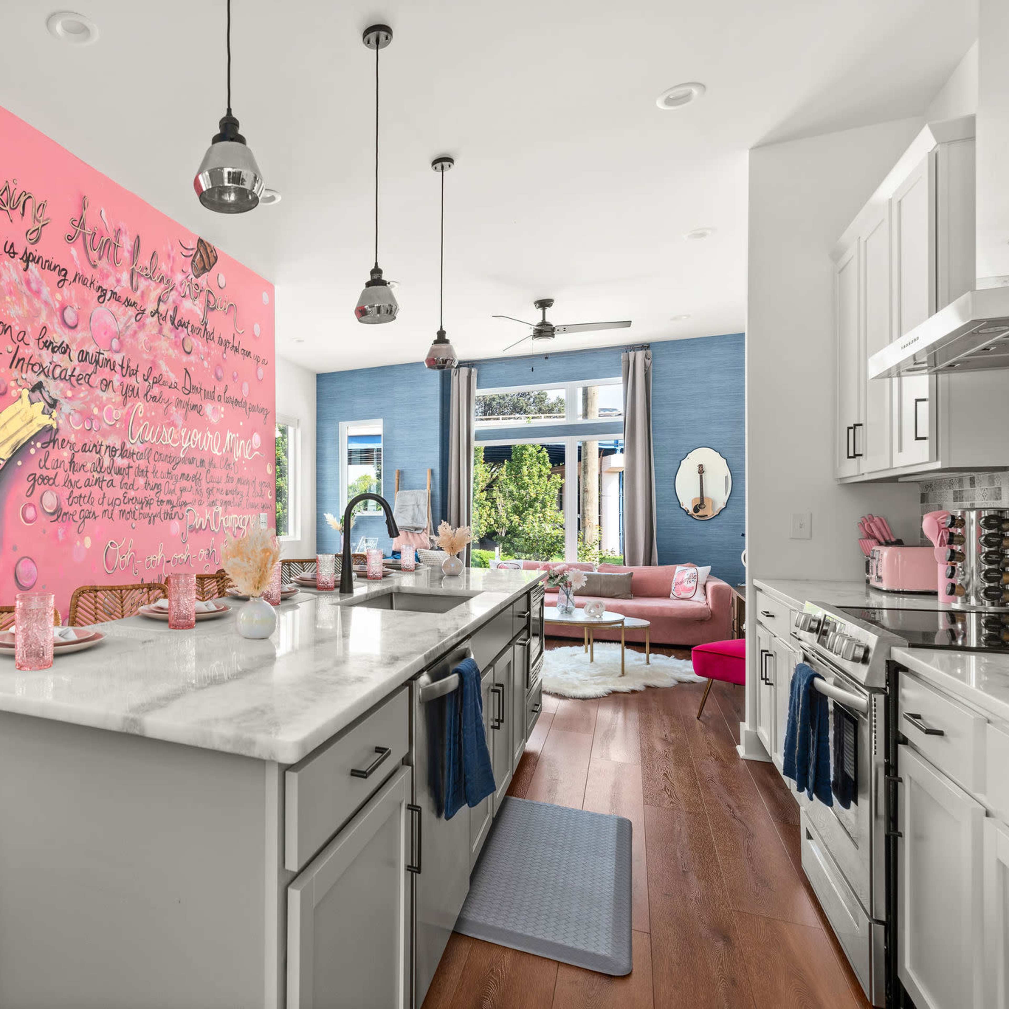 A modern kitchen features a pink mural of a wine bottle, with a marble countertop and a view into a brightly furnished living area.