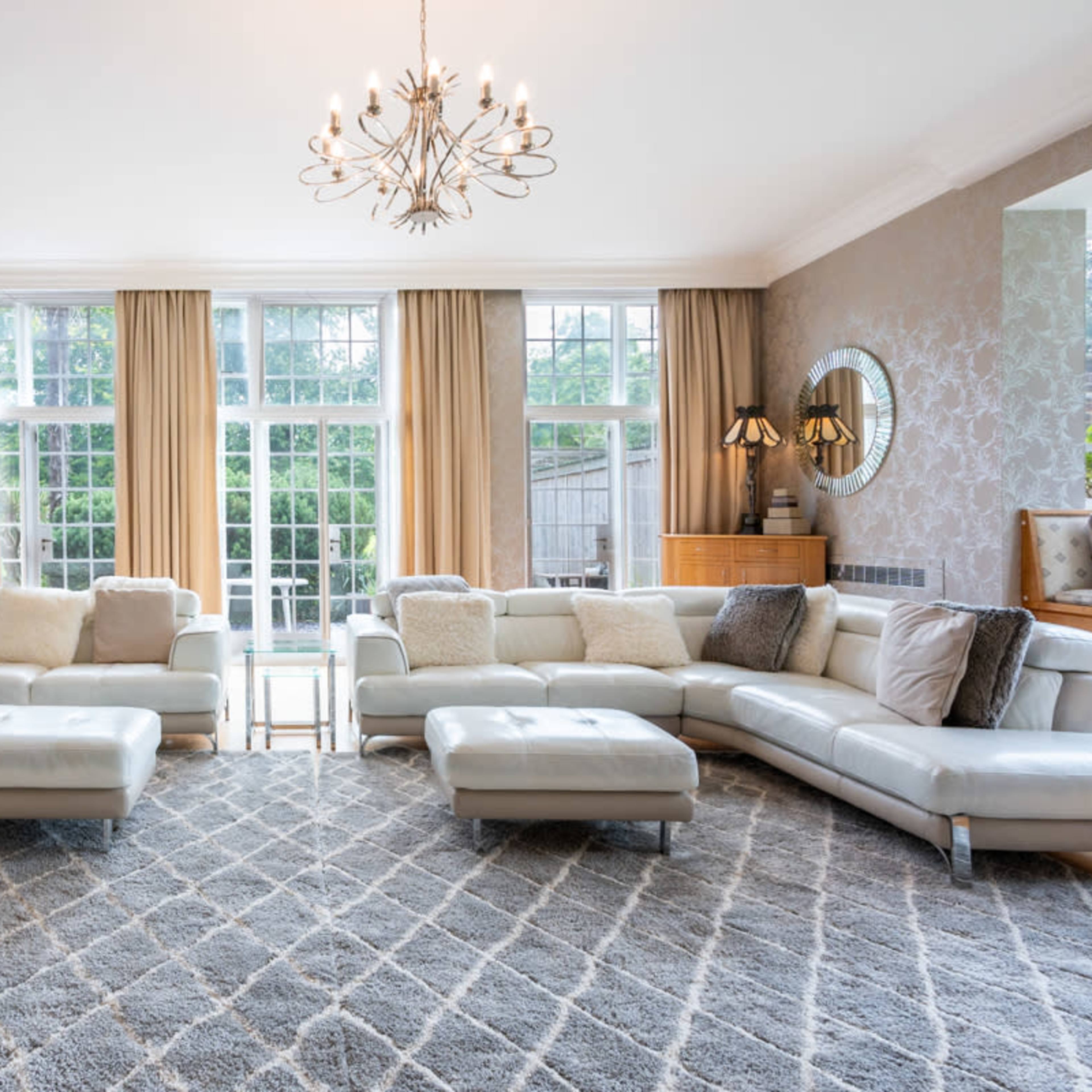 A spacious living room features a large white leather sectional sofa, a decorative chandelier, and large windows with curtains that provide ample natural light.