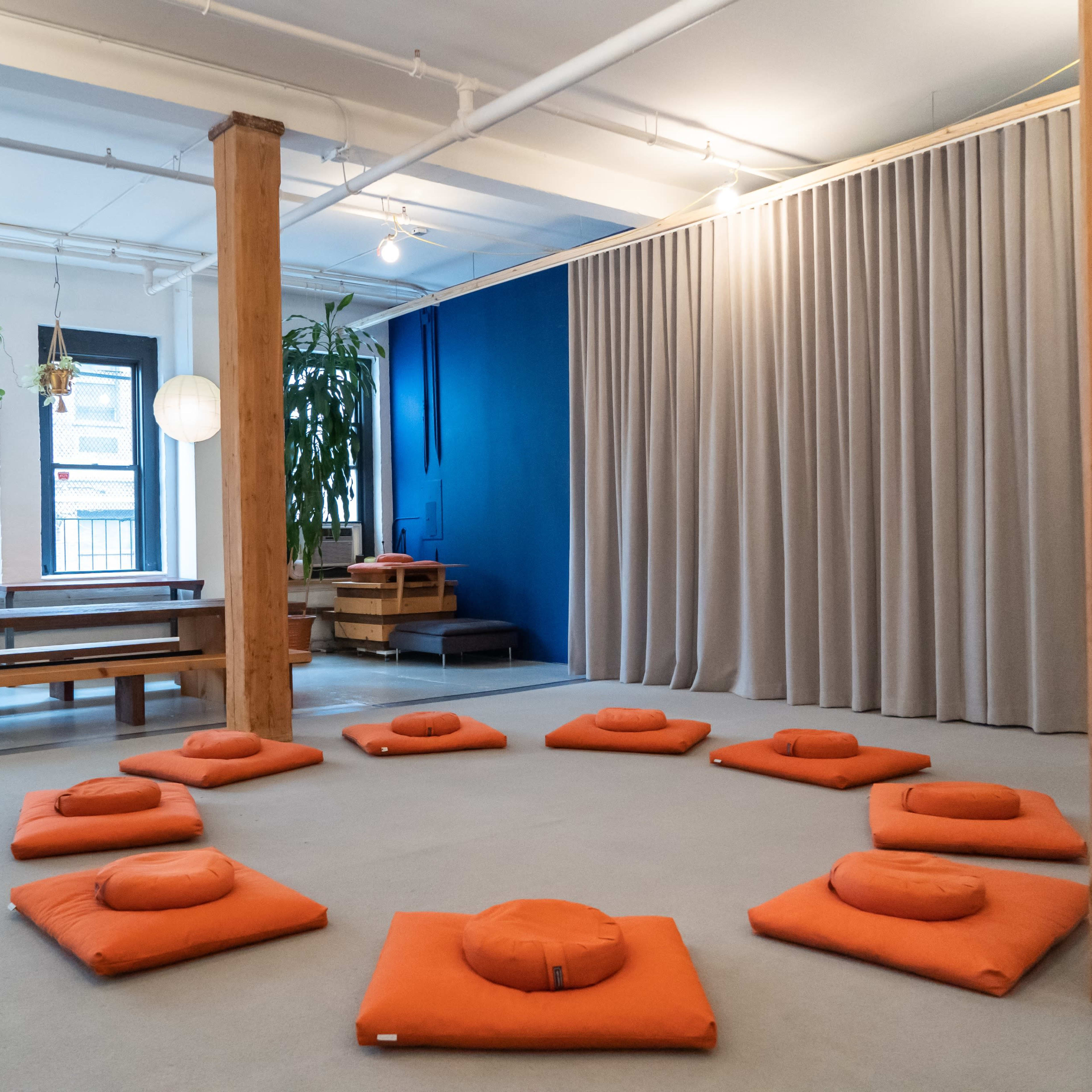 A meditation space with orange cushions arranged in a circular formation on a light-colored floor, surrounded by wooden beams and a curtain.