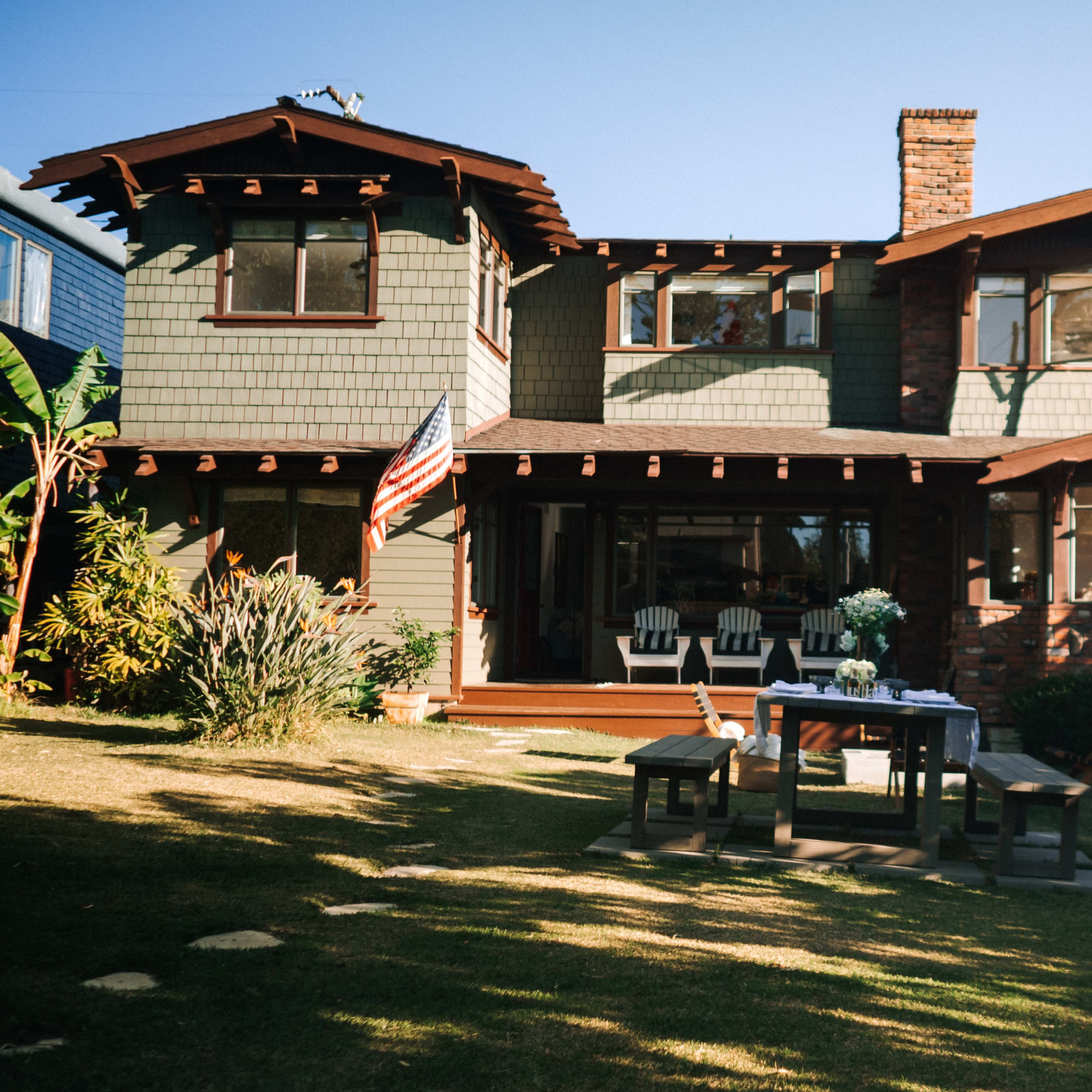 A two-story house with green shingle siding and wooden trim features a front porch with white chairs and a table in the yard, surrounded by shrubs and trees.