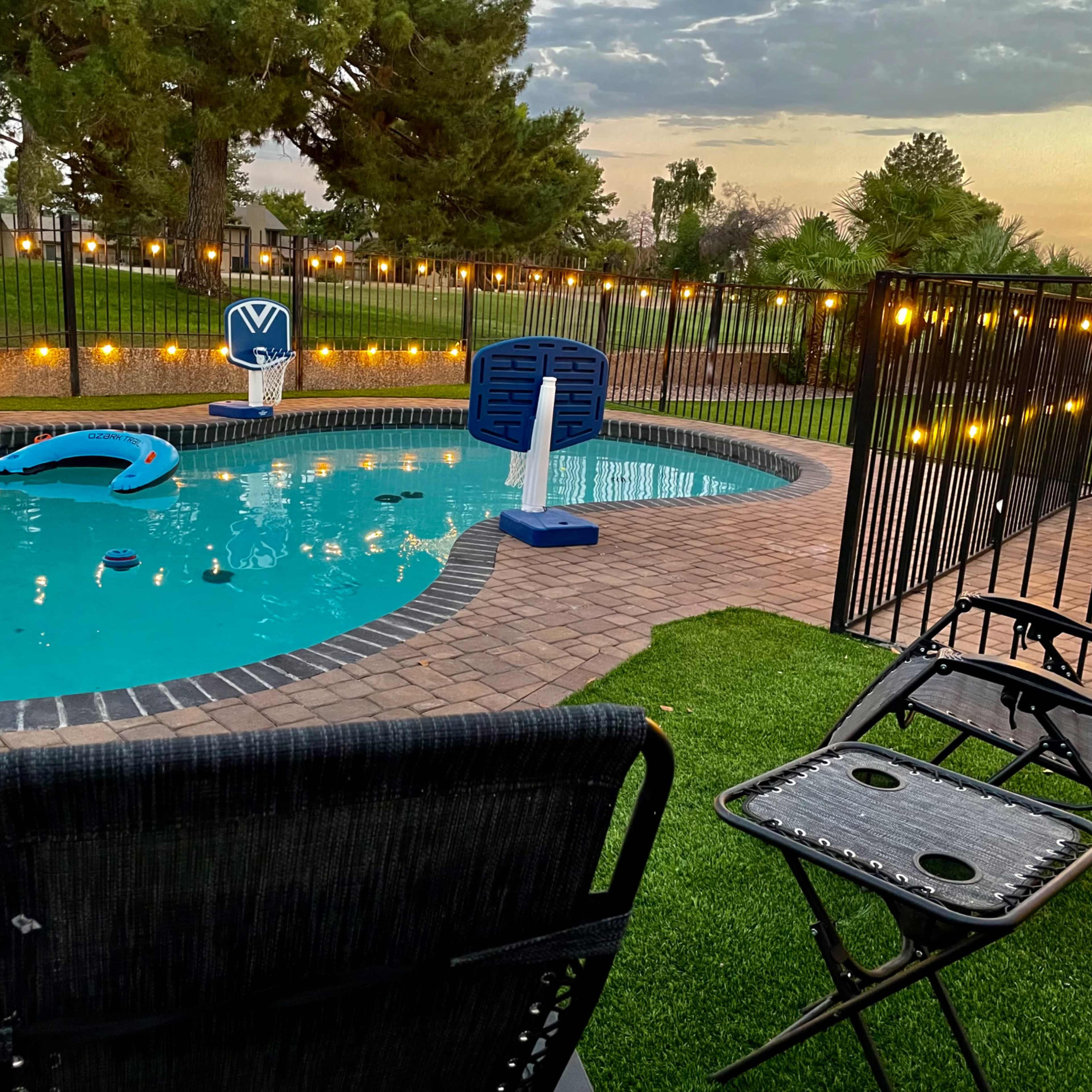 A swimming pool is surrounded by a paved patio, chairs, and decorative string lights, with grass and trees in the background.