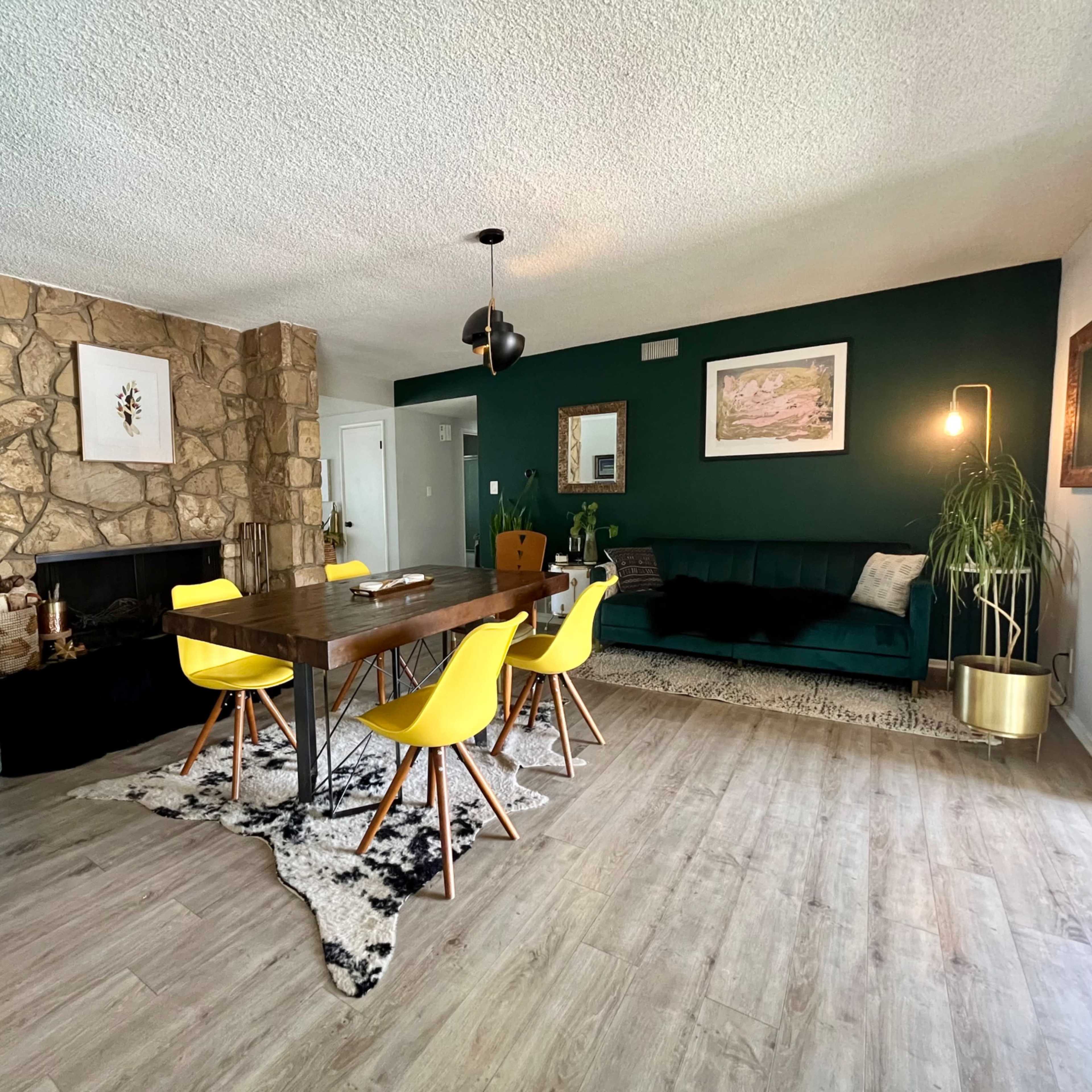 A modern living and dining area with a stone fireplace, a wooden dining table surrounded by yellow chairs, and a dark green accent wall.