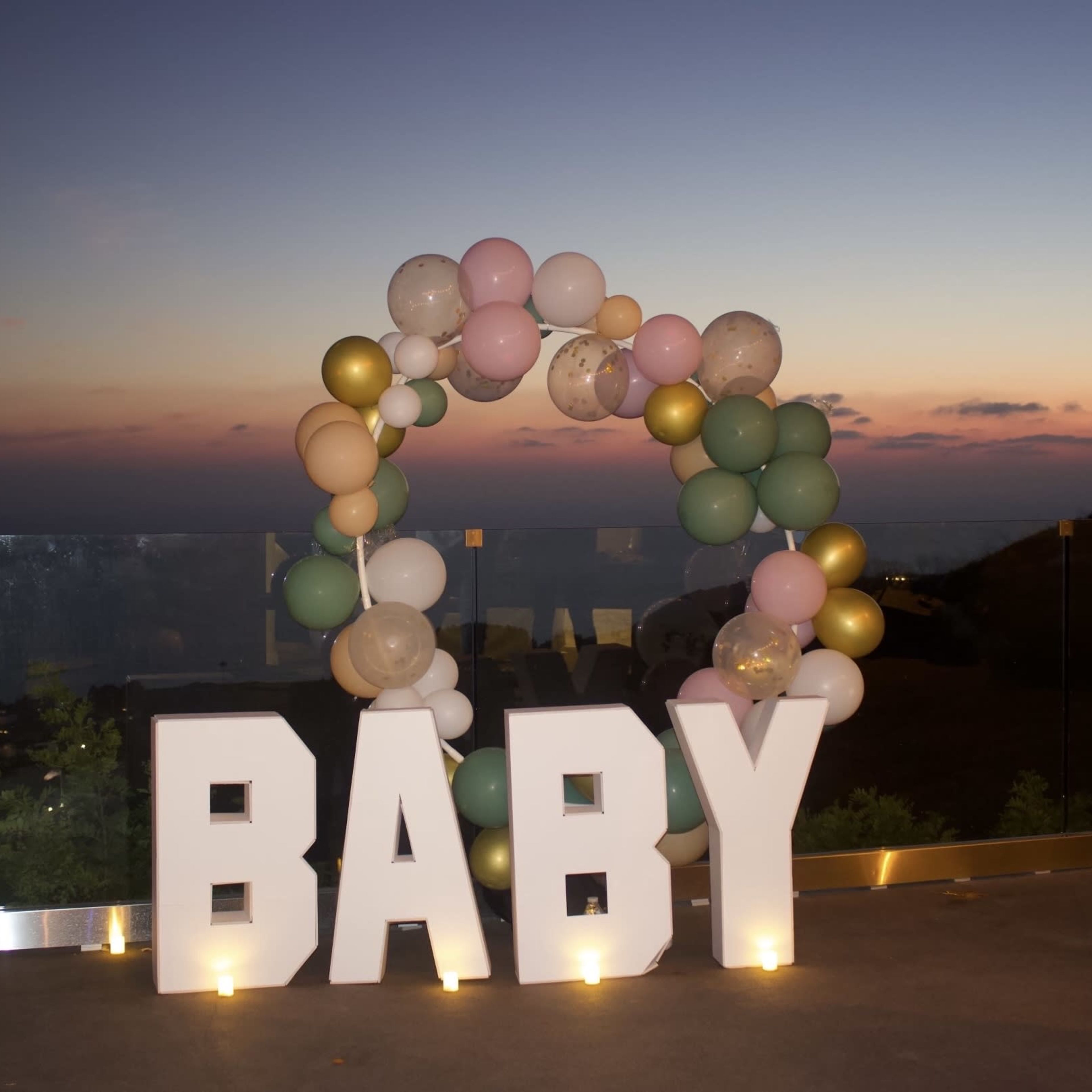 A decorative arch made of balloons surrounds the word "BABY" in large white letters against a sunset backdrop.