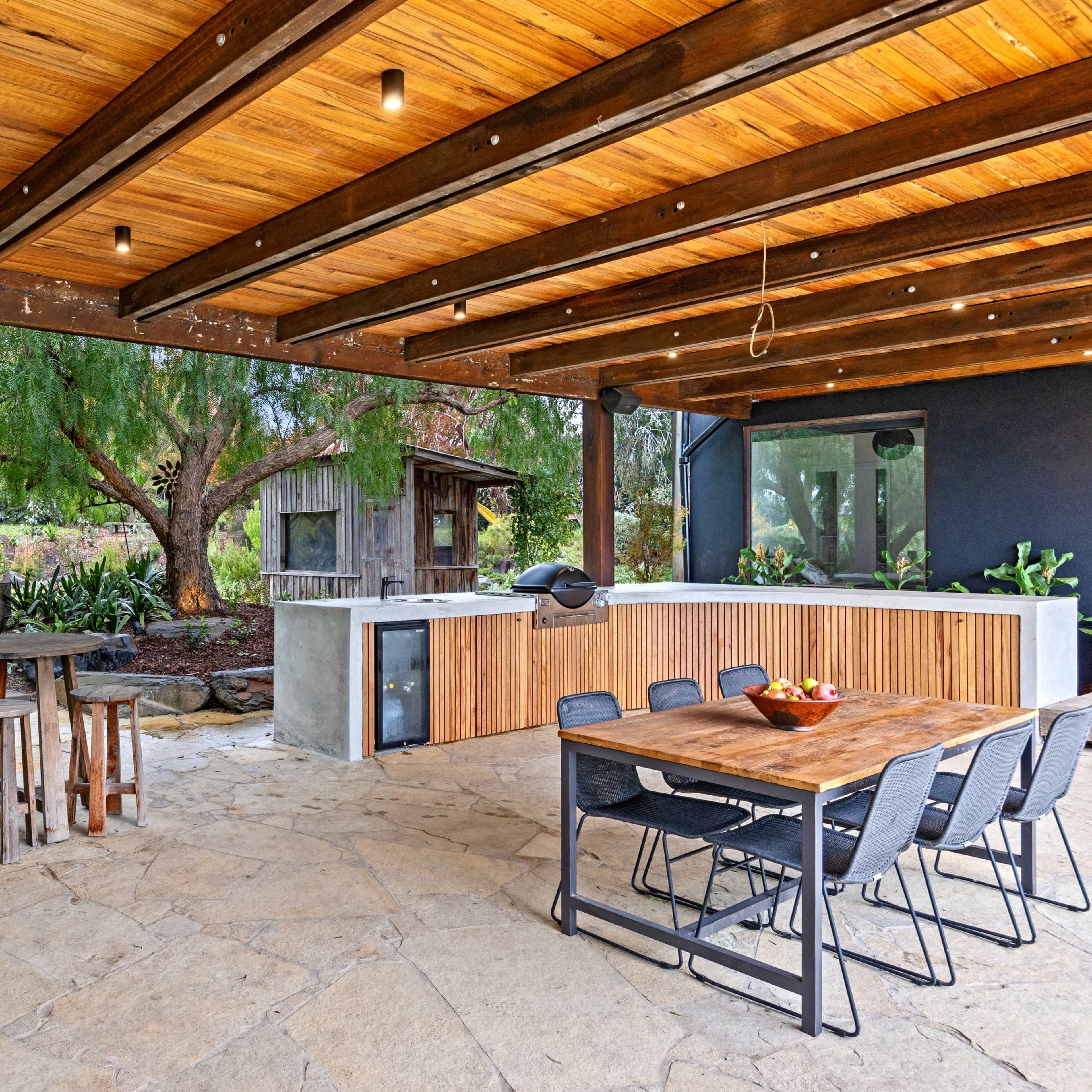 A covered outdoor patio features a wooden dining table and chairs, a kitchen area, and a stone floor, surrounded by greenery and trees.