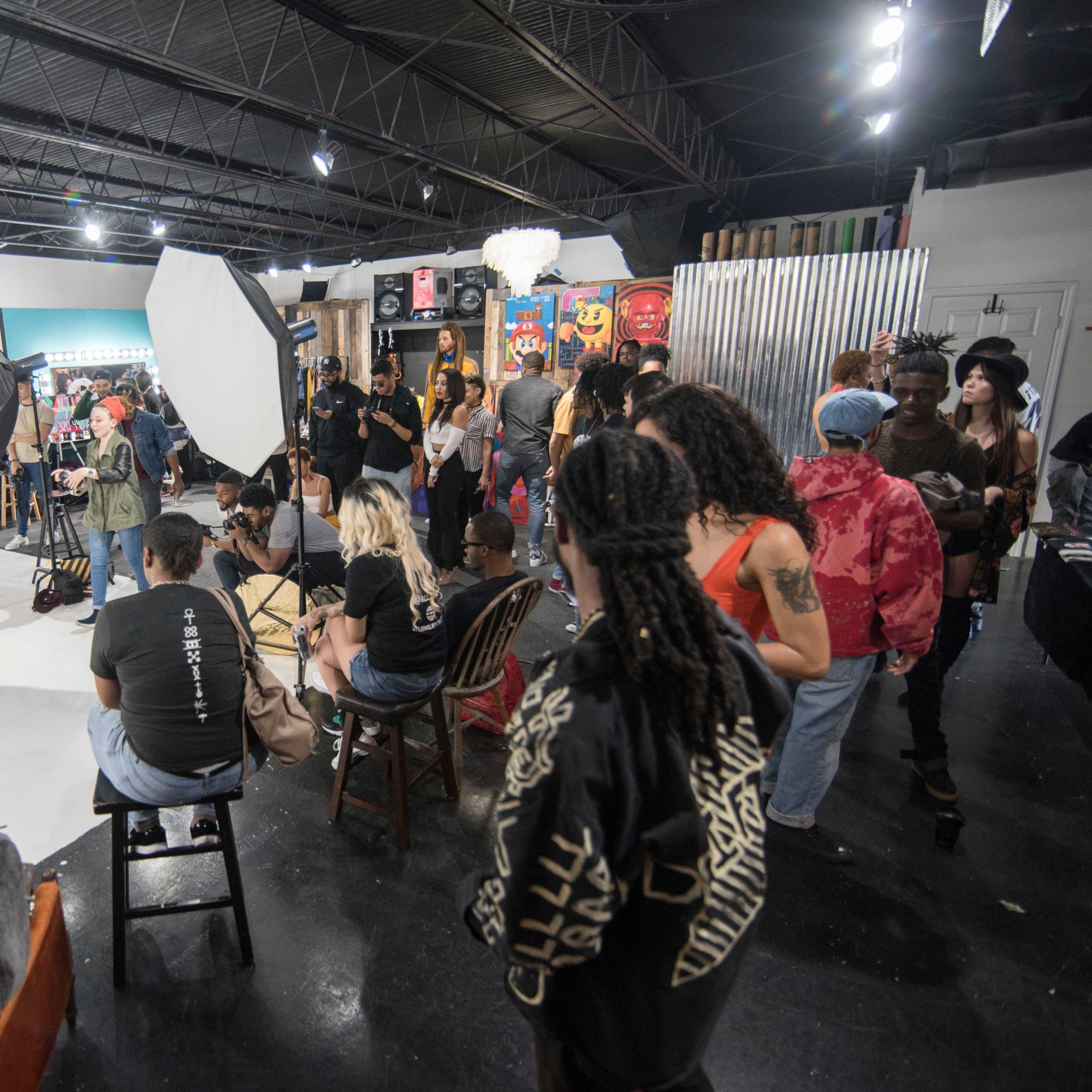 A group of people observes a photo shoot taking place in a studio with a white backdrop and various props.