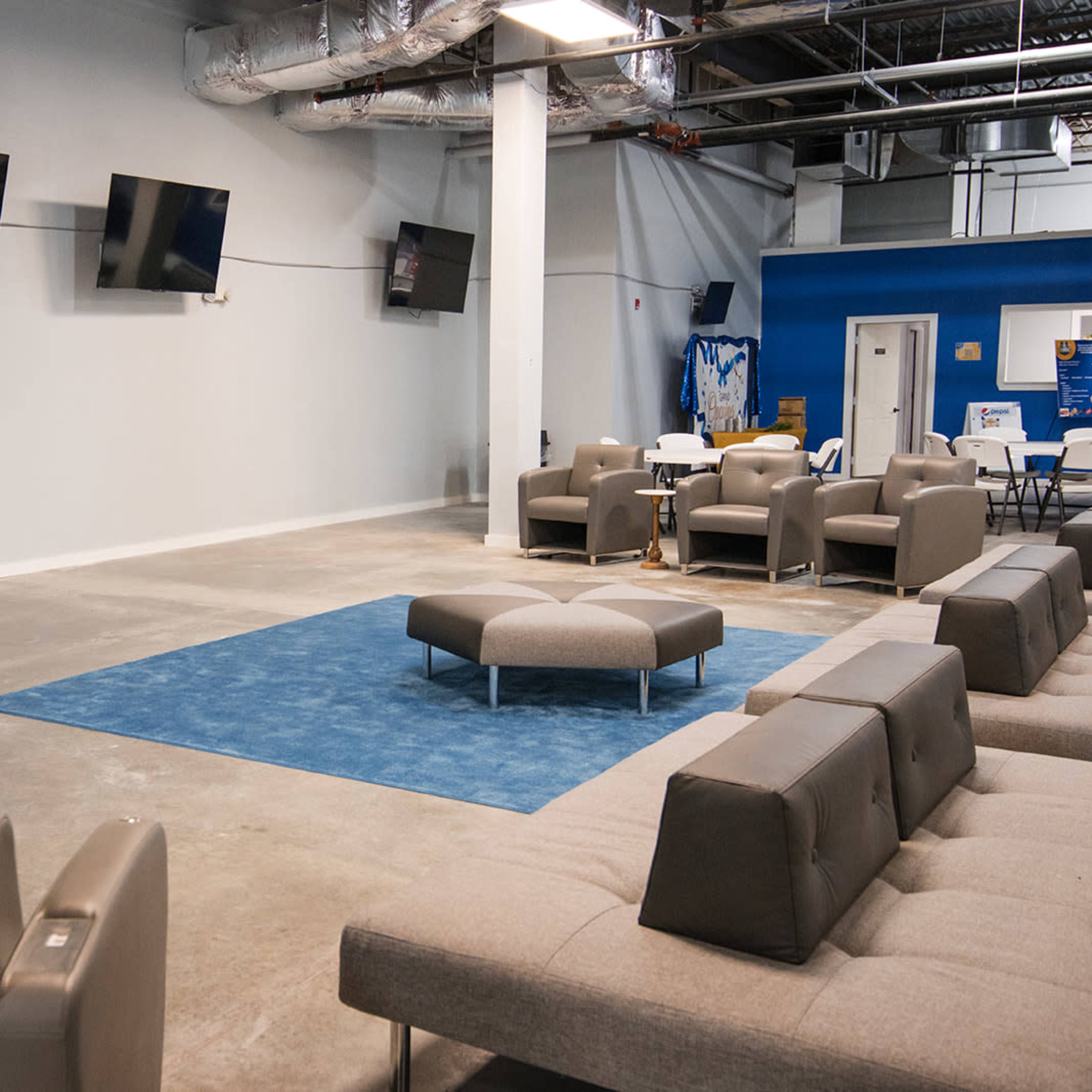 A spacious lounge area with several wall-mounted televisions, grey seating arrangements, and a large blue rug on a concrete floor.