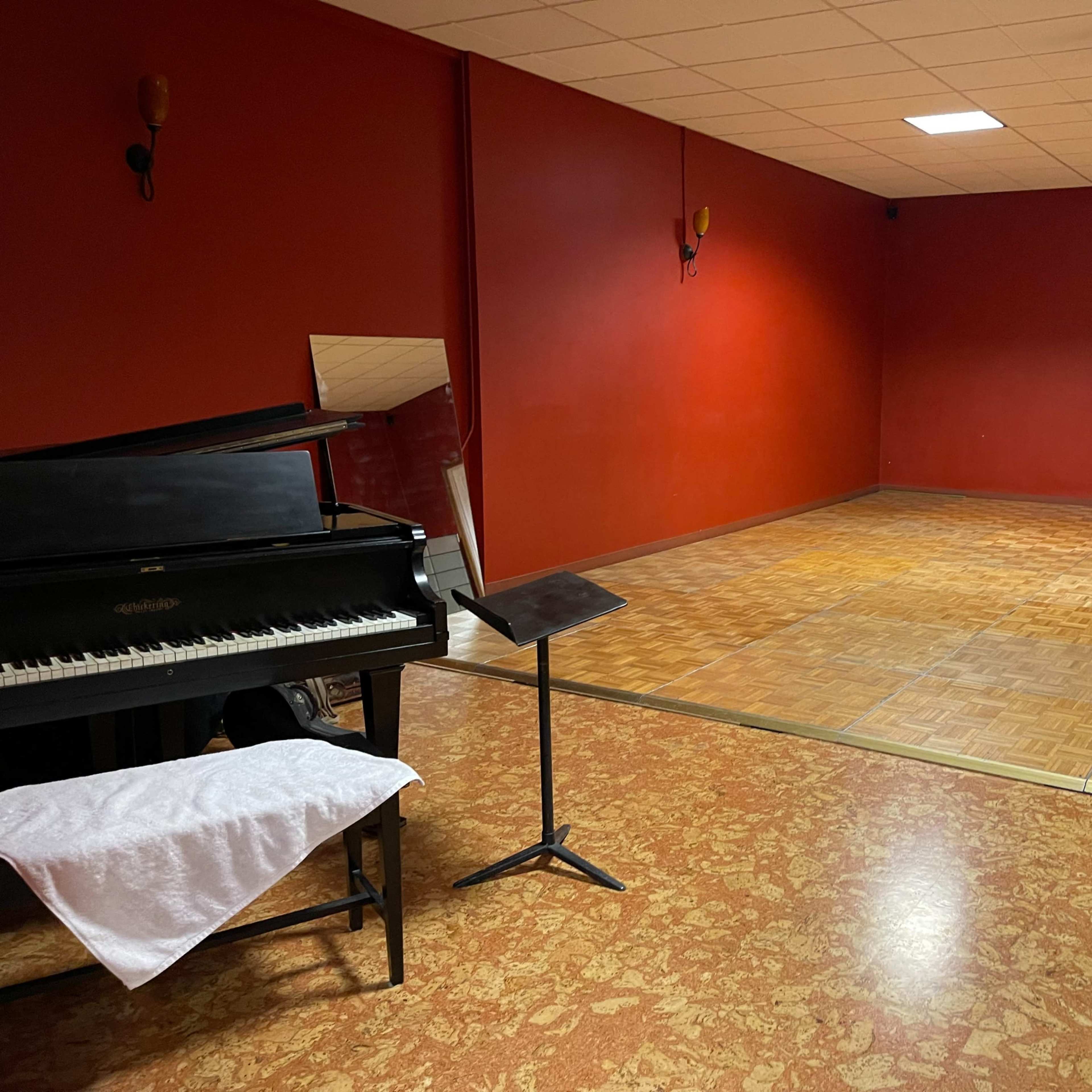 A room with red walls featuring a black piano, a music stand, and a wooden dance floor area.
