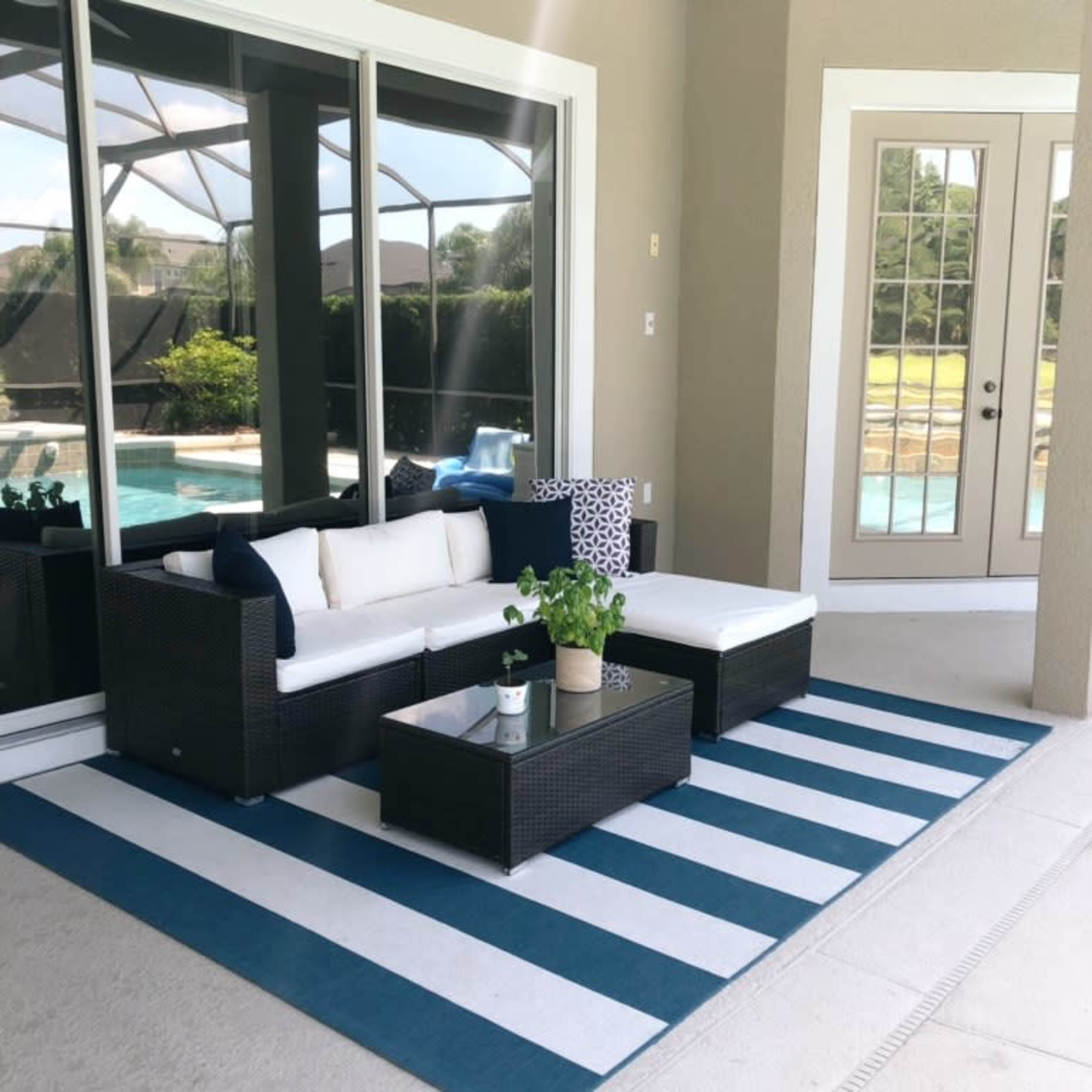 A patio area with a white and blue striped rug, a wicker seating arrangement, and a small table with a plant, all facing a large window overlooking a pool.