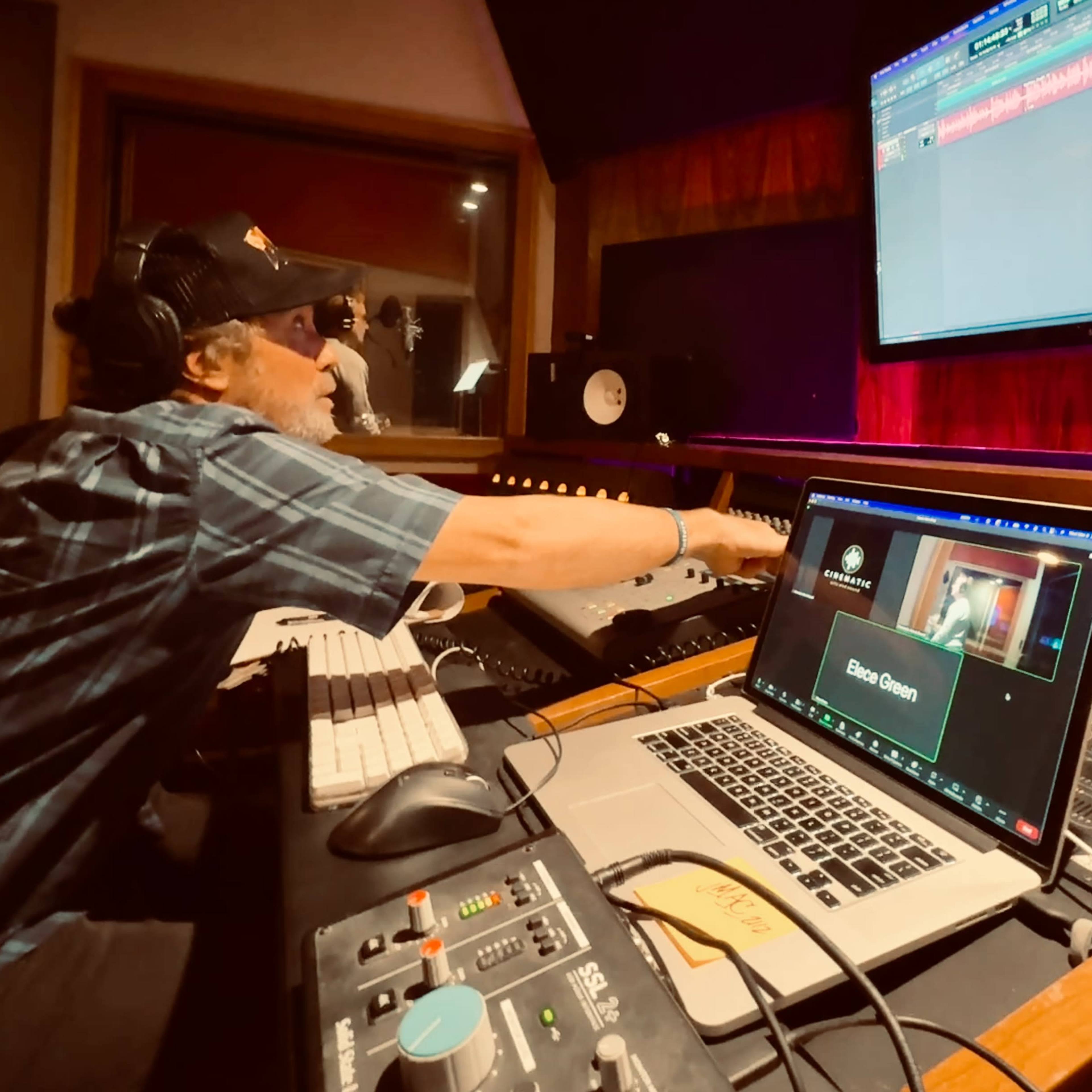 A person is working in a music studio, adjusting controls on a console while looking at a computer screen displaying audio tracks.
