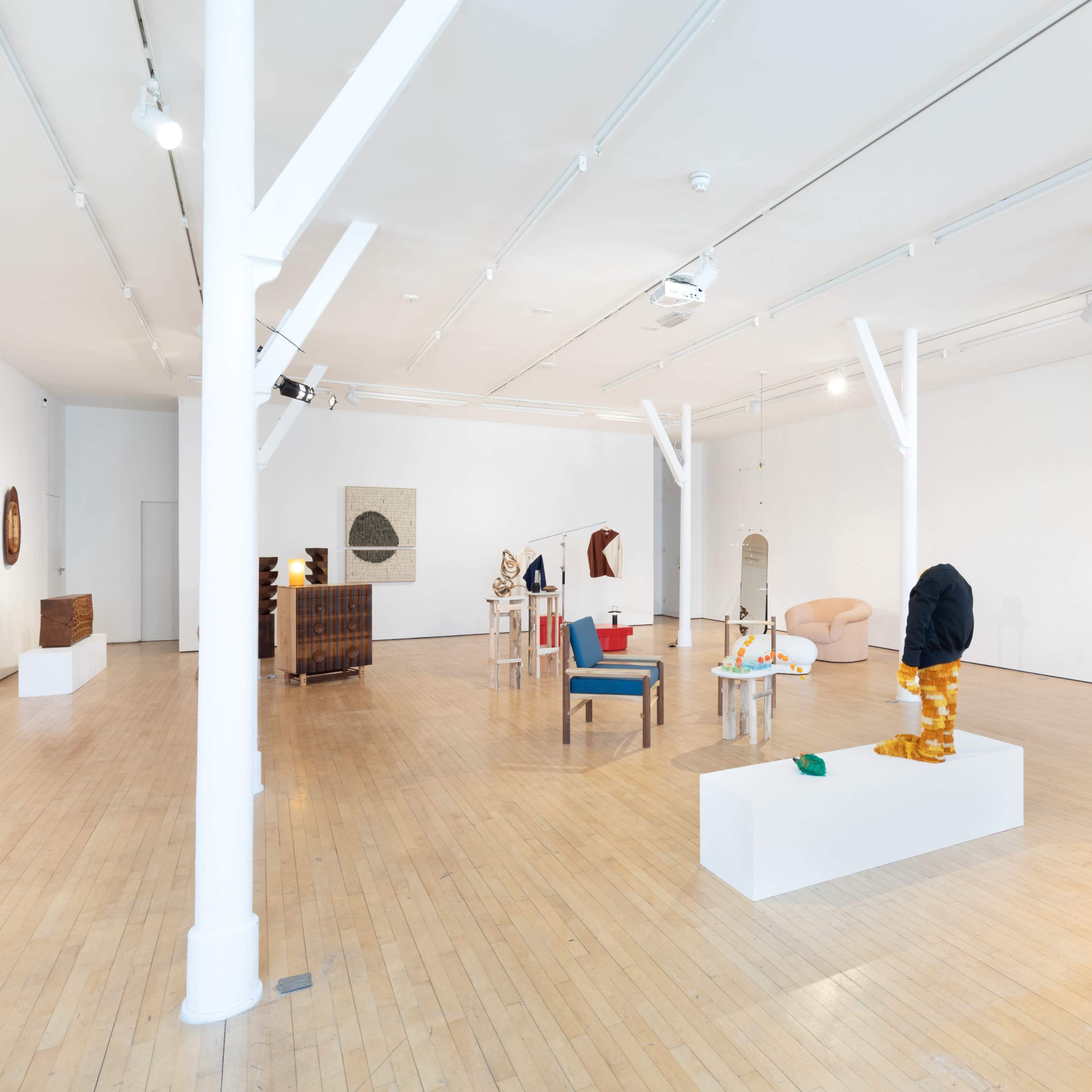 A spacious art gallery with wooden floors and white walls, featuring various sculptures and artworks displayed on pedestals and furniture throughout the room.