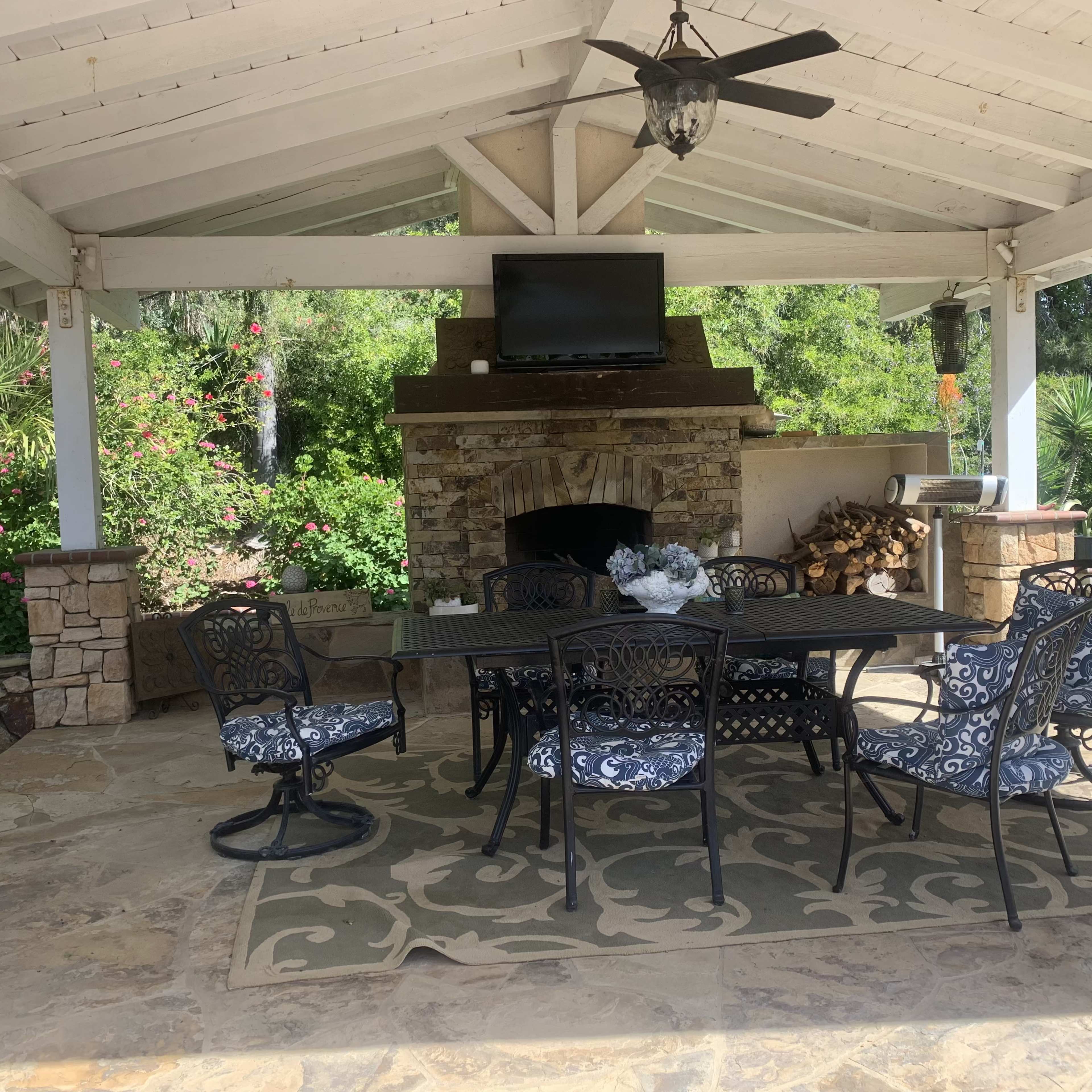 A covered outdoor patio features a large dining table surrounded by black metal chairs, with a stone fireplace and a television mounted on the wall.
