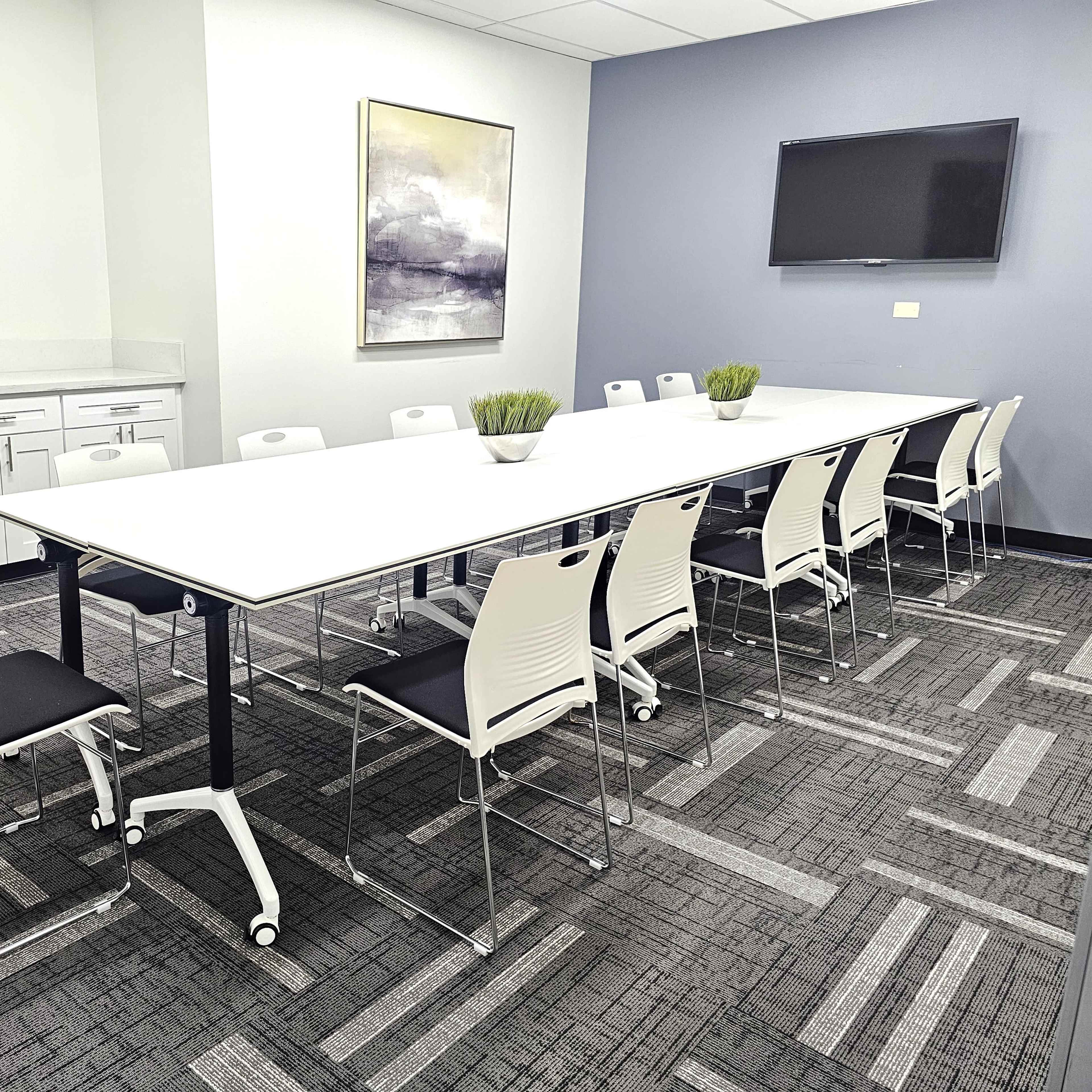 A long, white conference table is set up in a meeting room surrounded by twelve chairs, with a flat-screen TV on the wall and a side cabinet in the background.
