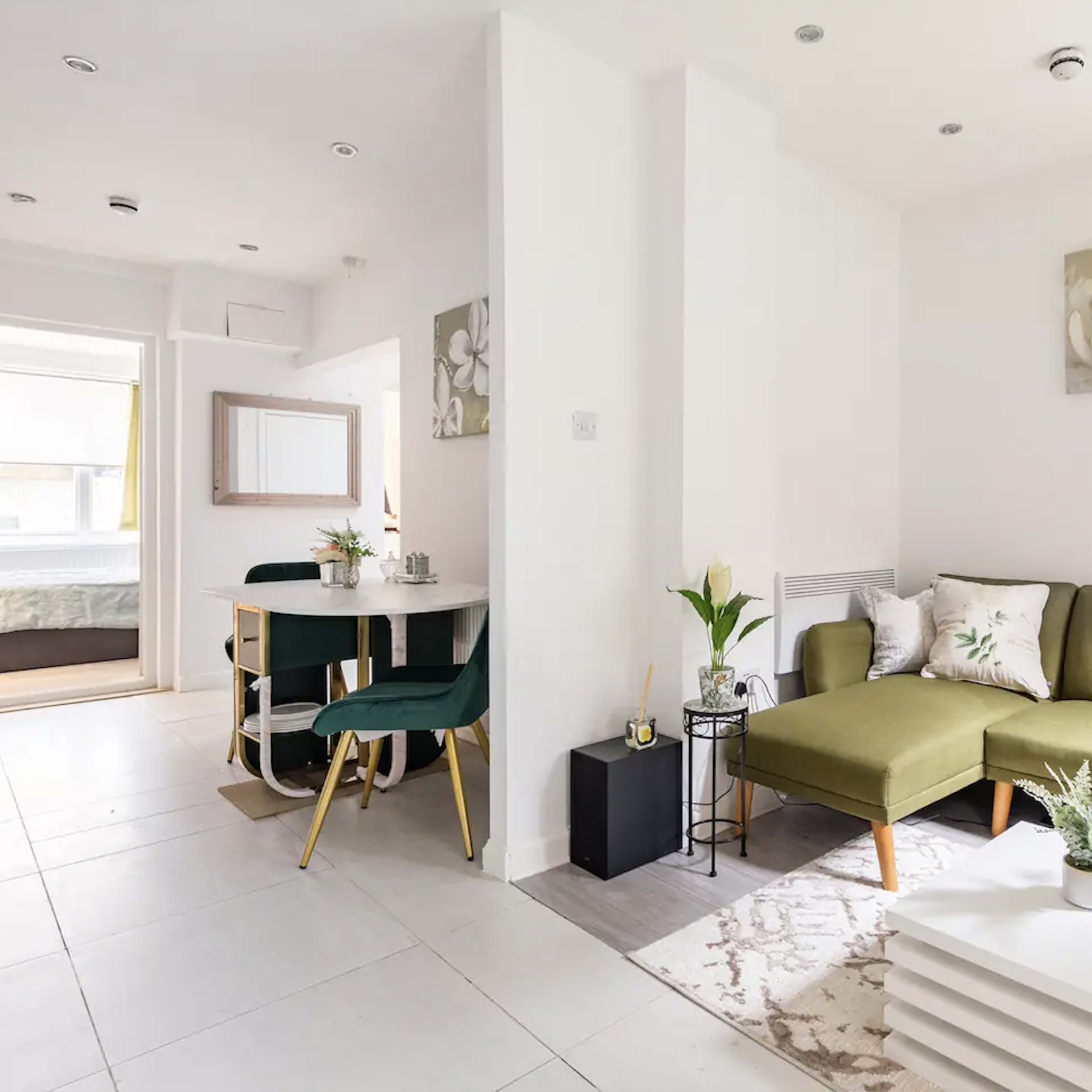 A modern living area features a green sectional sofa, a white coffee table, and a dining space with a round table and chairs, all set against a light-colored wall.