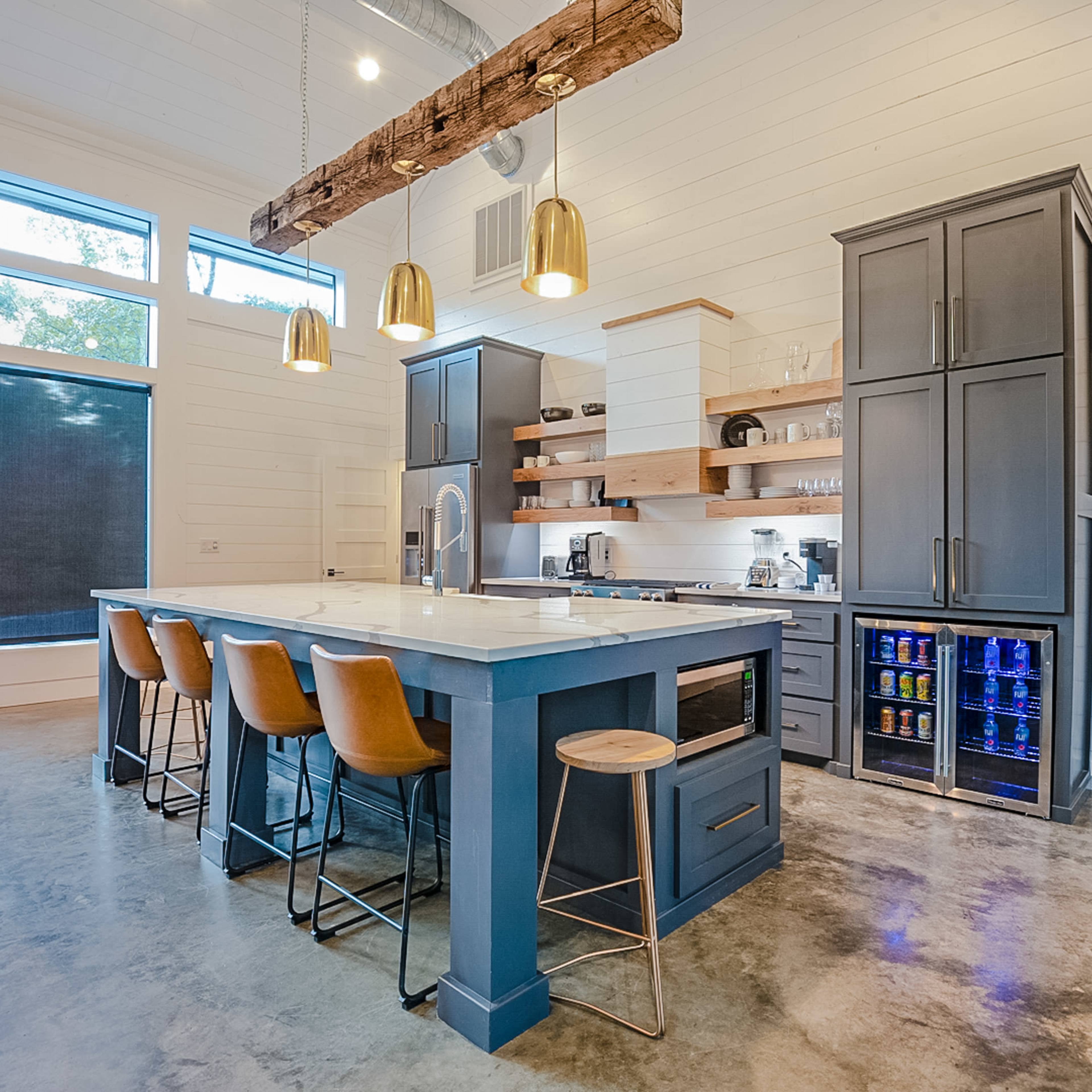 A modern kitchen with a large island, wooden bar stools, and a combination of gray cabinetry and white walls, illuminated by pendant lights.
