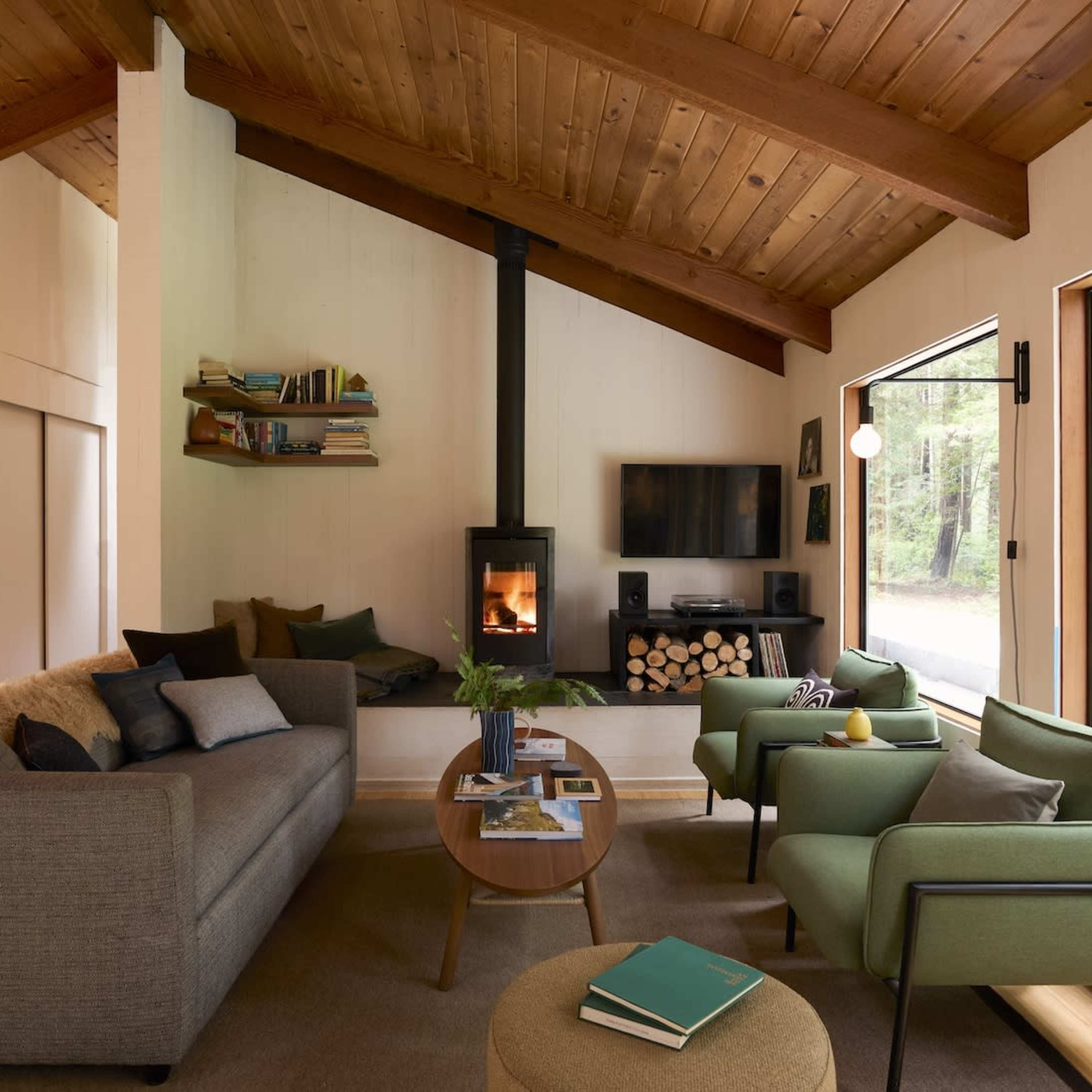 A modern living room with a gray sofa, two green chairs, a wooden table, a fireplace, and large windows overlooking a forest.